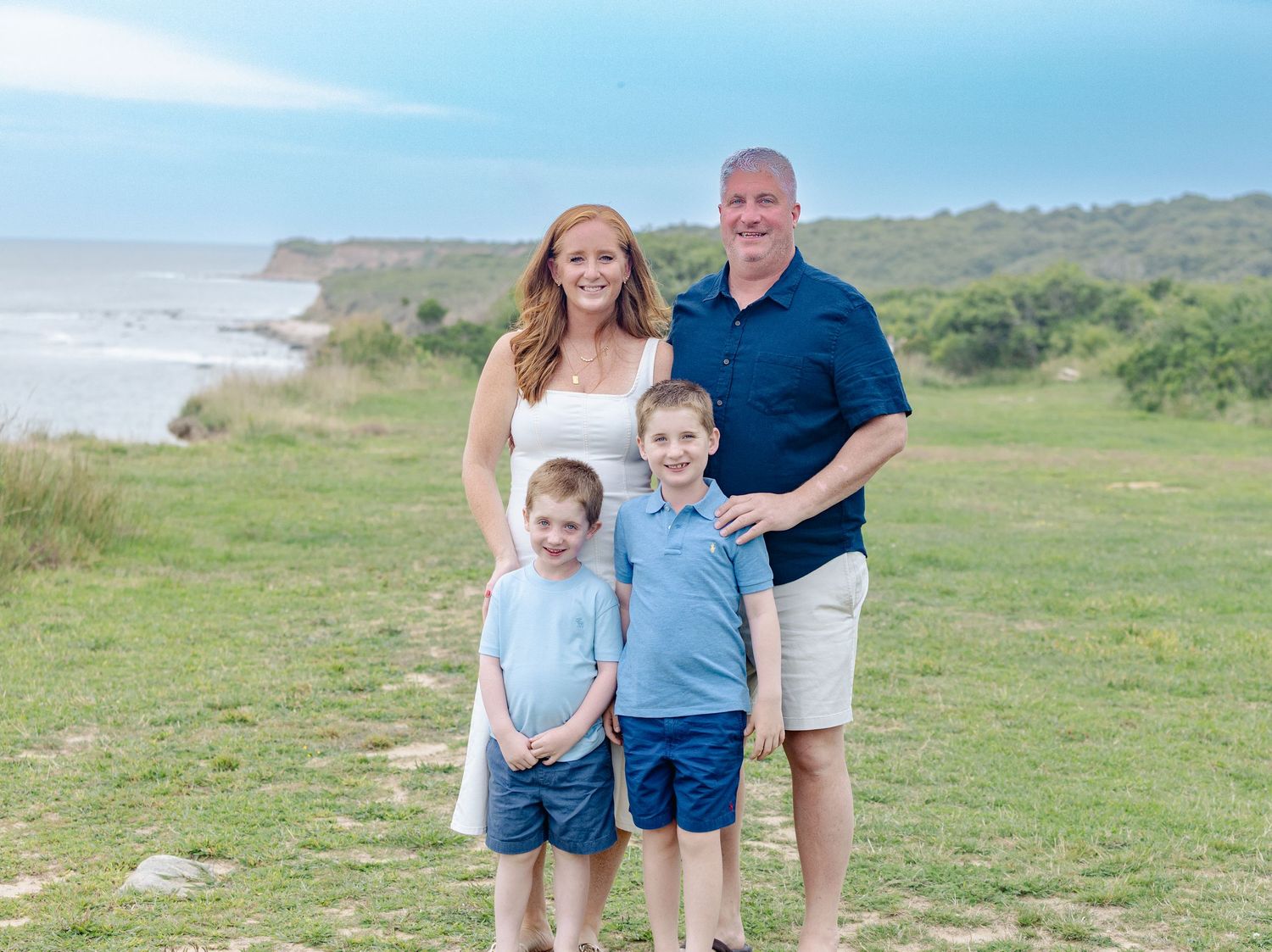 Family of four smiling outdoors on green coastal cliffs with ocean views on a cloudy day.