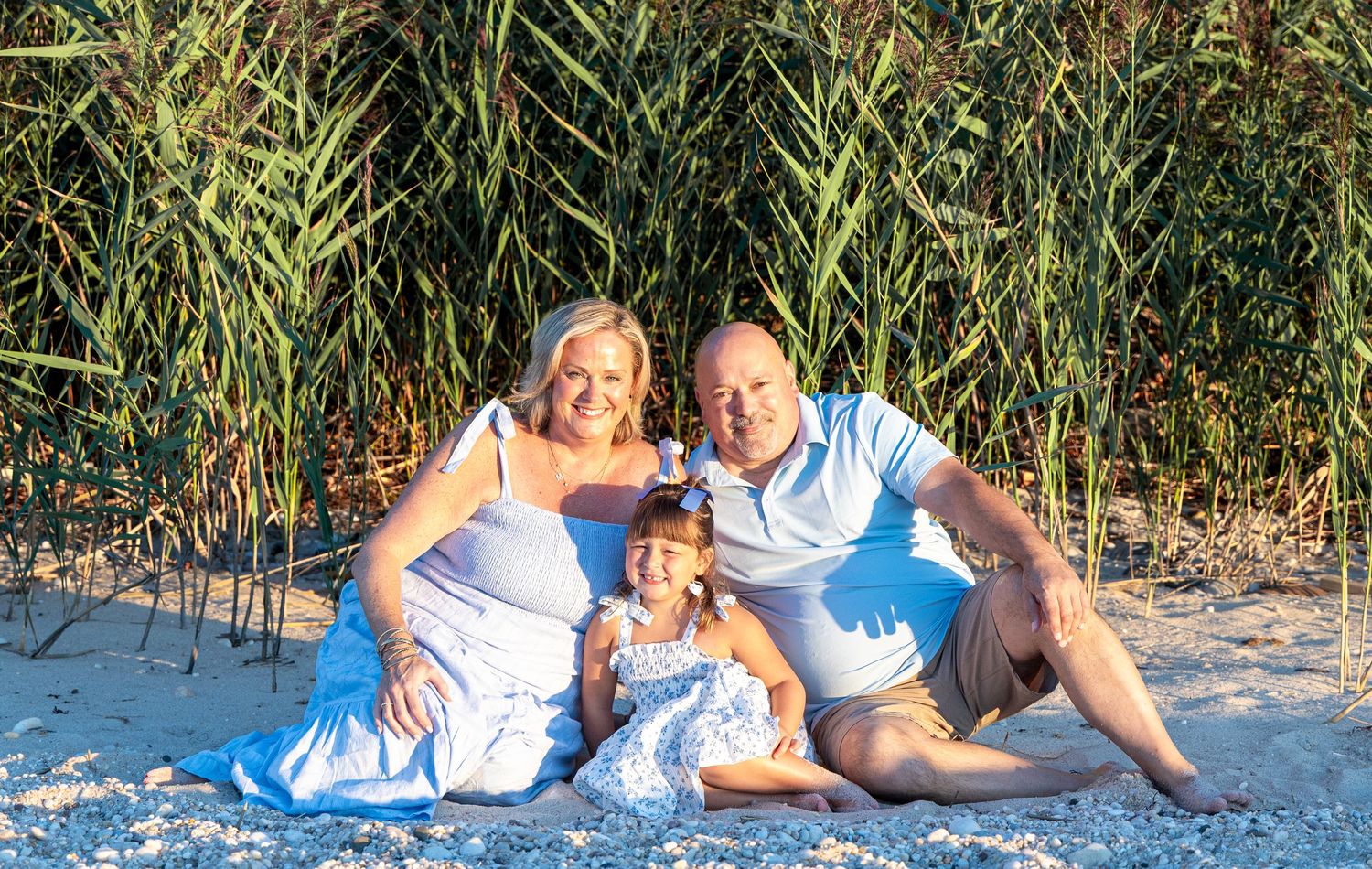 Family of three smiling on sandy beach with lush green reeds, parents in white outfits with young daughter in blue dress. north fork. beach