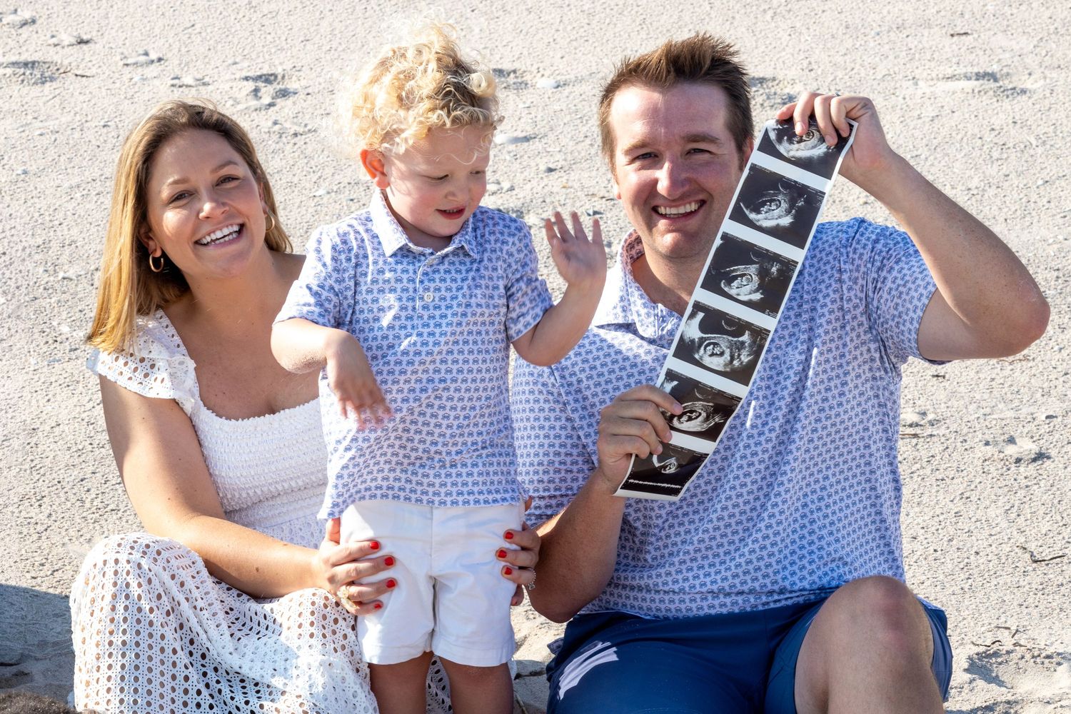 Happy family on beach holding ultrasound photos, pregnancy announcement with toddler son.
