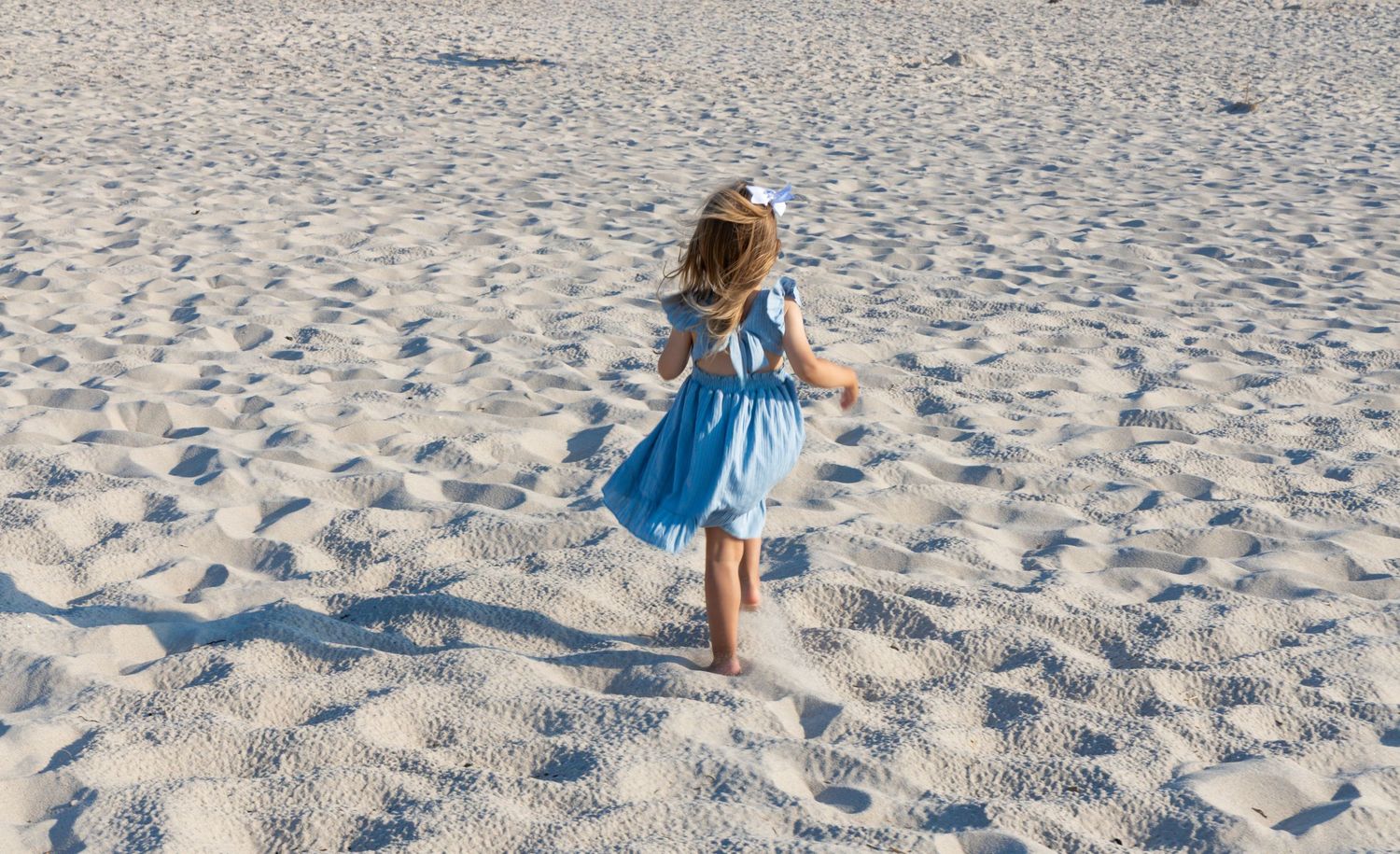 Young girl in blue dress running barefoot across white sandy beach.
