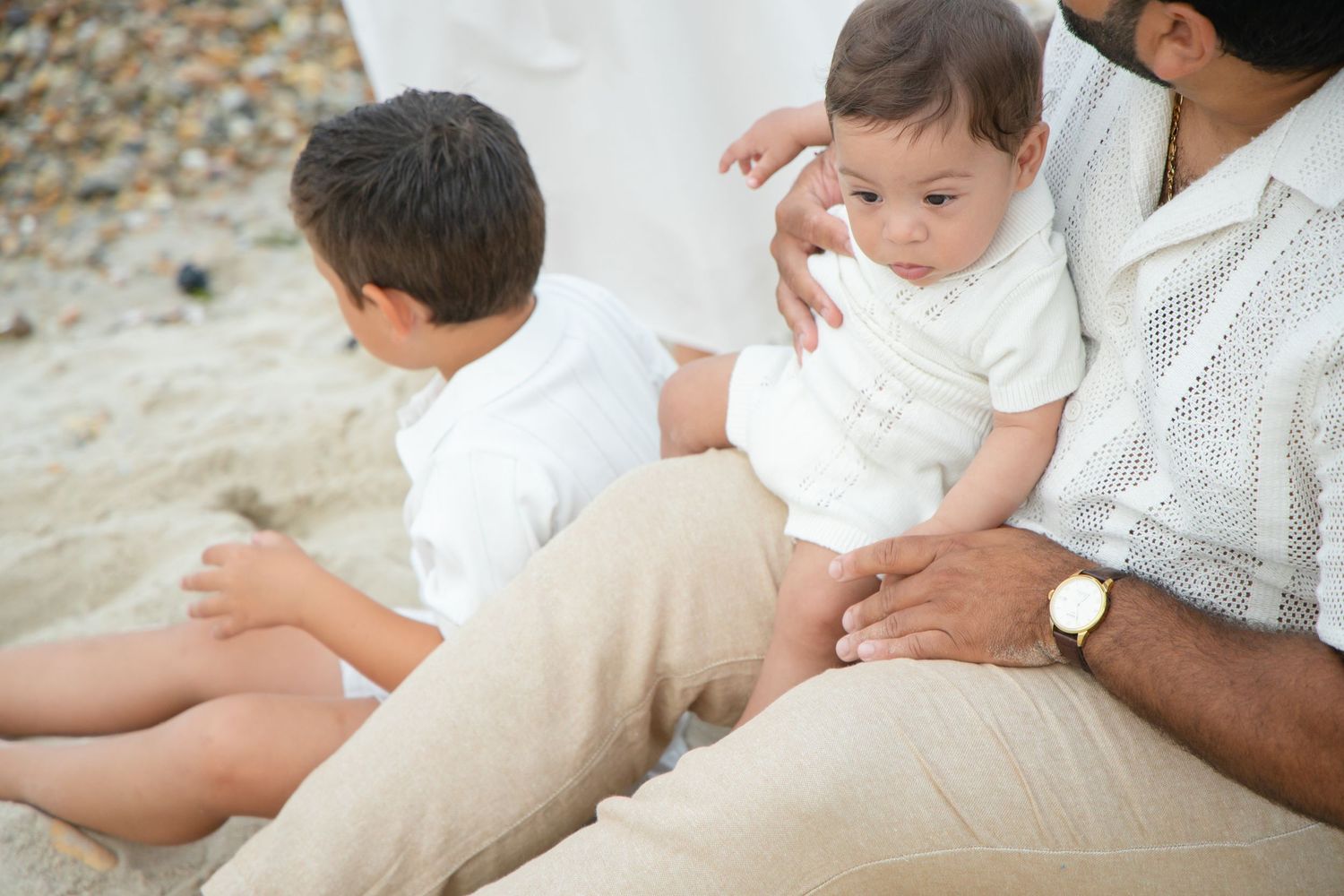 Father holding baby on beach with young boy sitting nearby, family beach portrait.