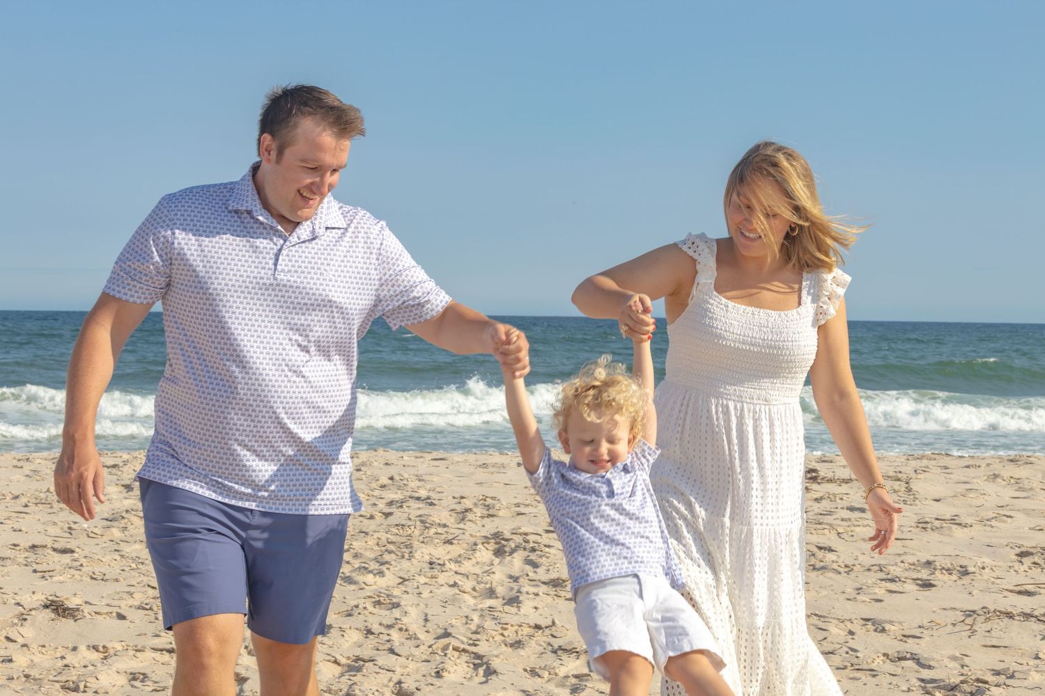 Family swinging young blonde child between parents while walking on sunny beach together.