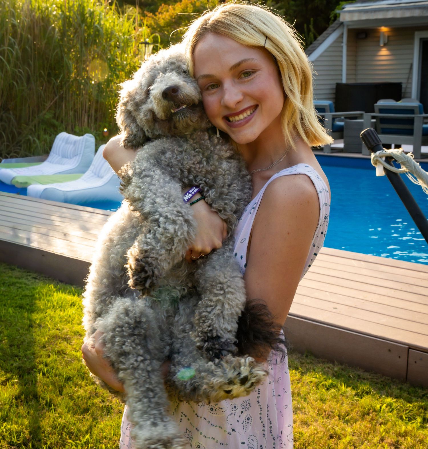 Smiling blonde woman hugging fluffy gray poodle puppy in backyard near swimming pool.