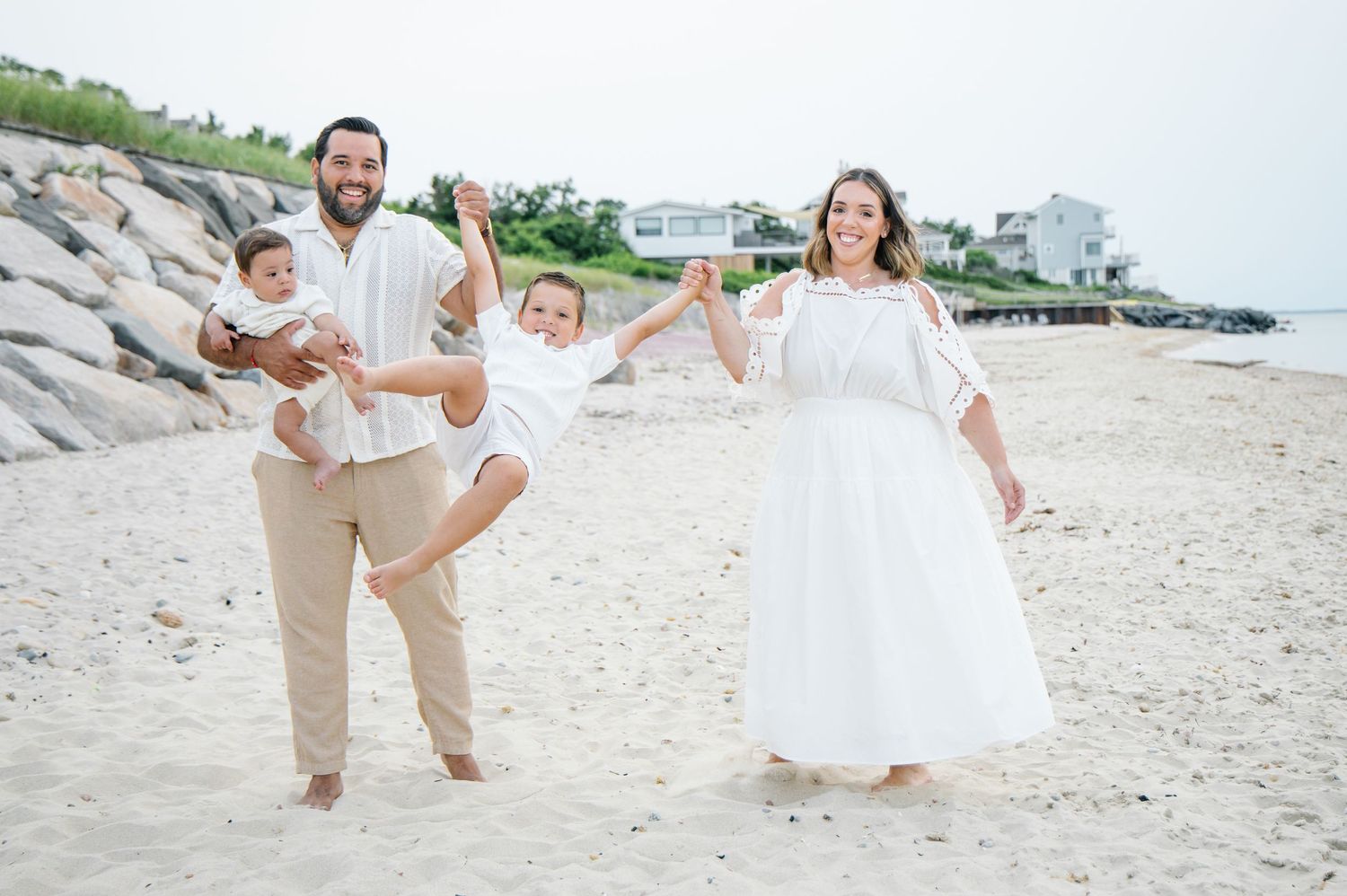Happy family of four in white outfits playing on a sandy beach, parents swinging children with joy.