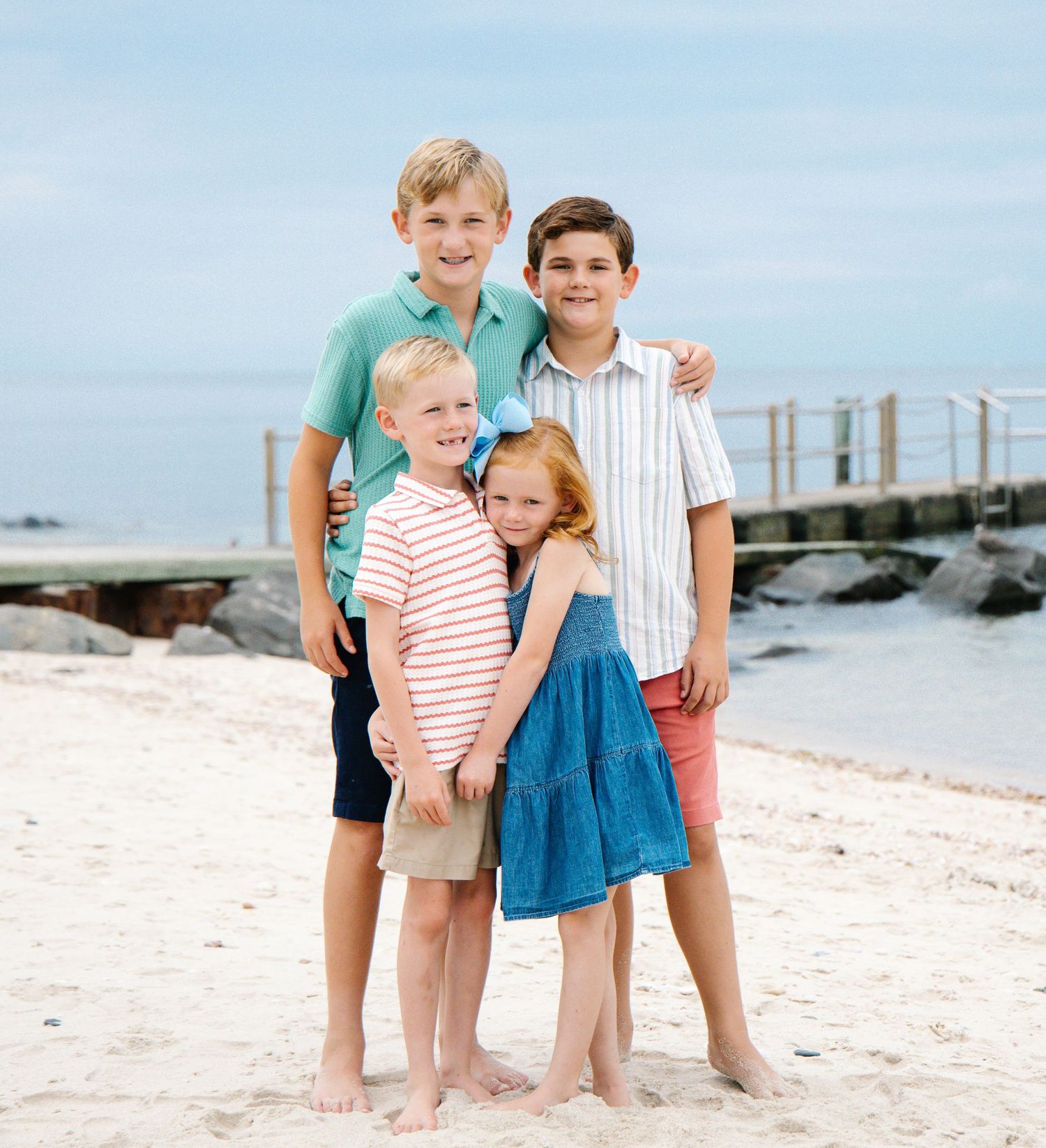 Four siblings posing together on sandy beach with rocky pier in the background.