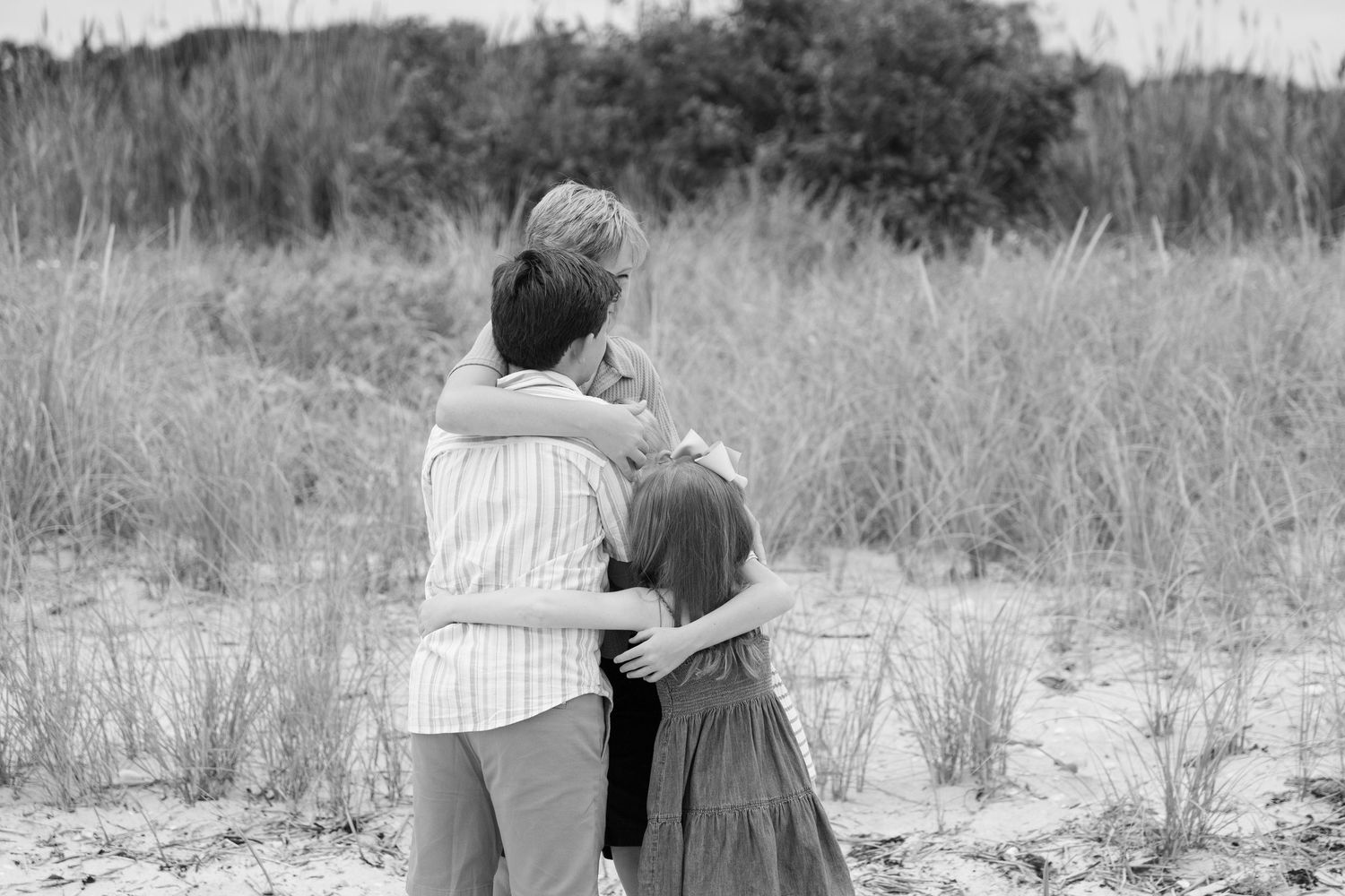 Black and white photo of woman hugging two children from behind near sandy dunes.