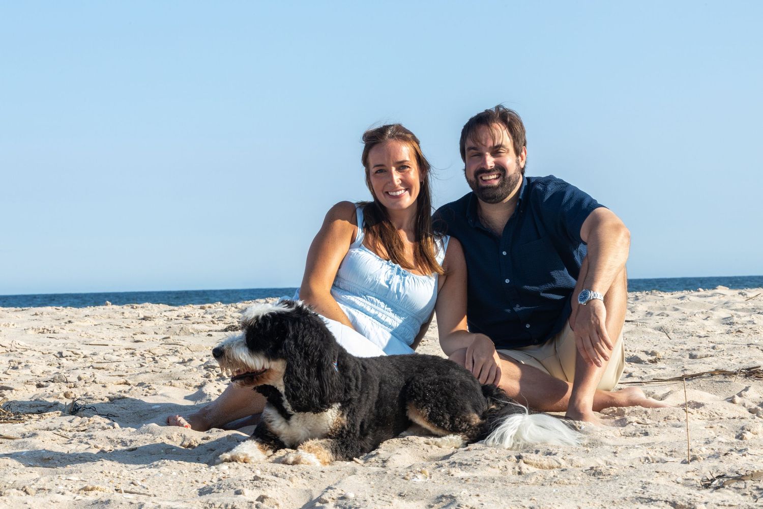 Smiling couple sitting on beach sand with their large black and white dog, blue sky behind them.