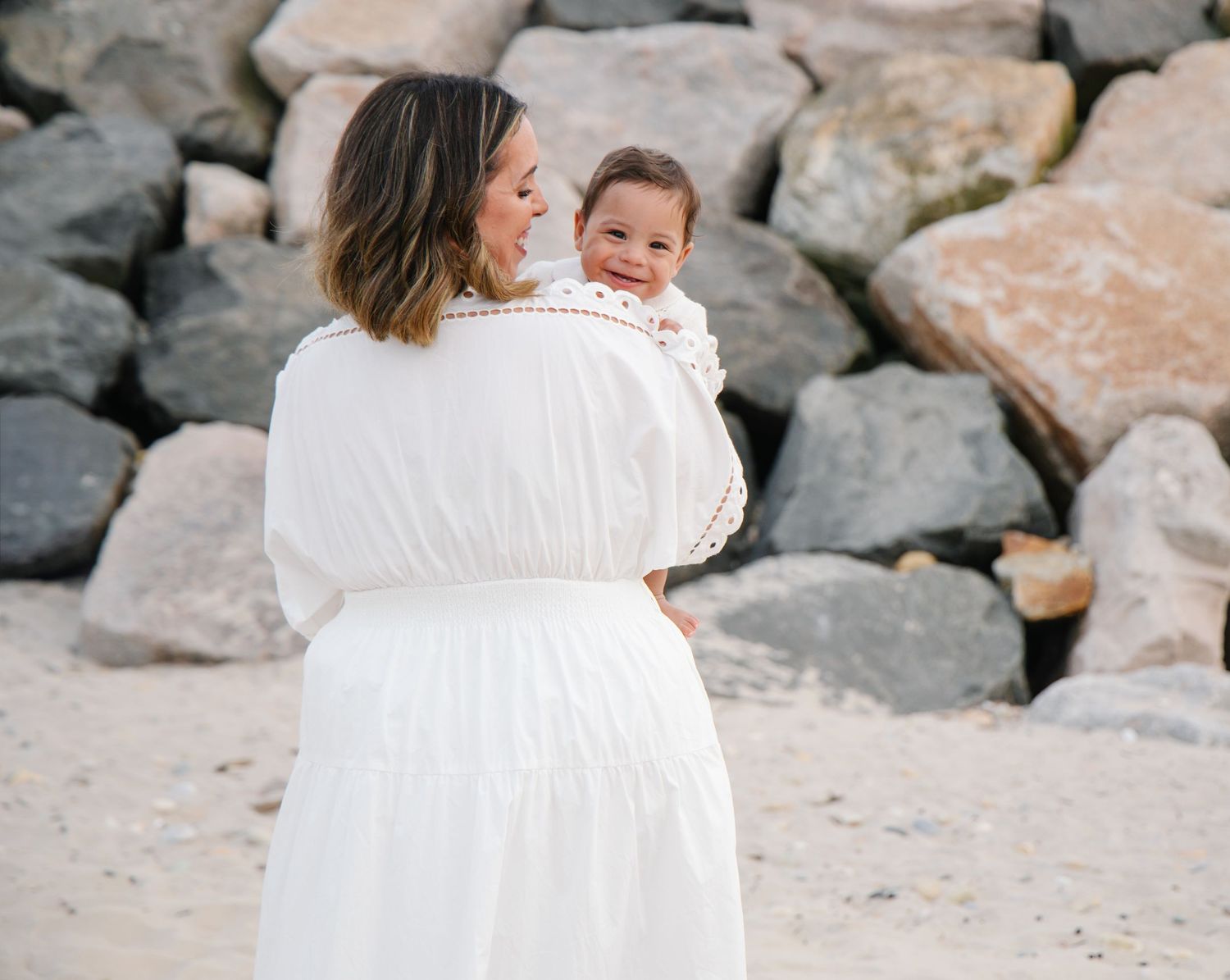 Mother in white dress holding smiling baby on sandy beach with large rocks behind.