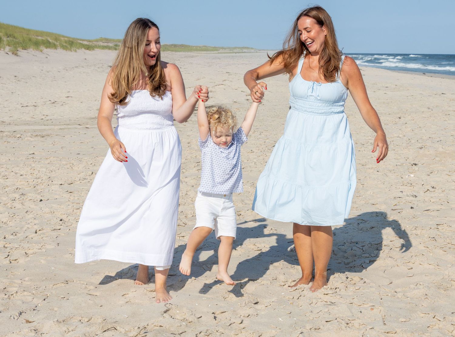 Two women in white and blue dresses swinging young child between them while walking on sunny beach.