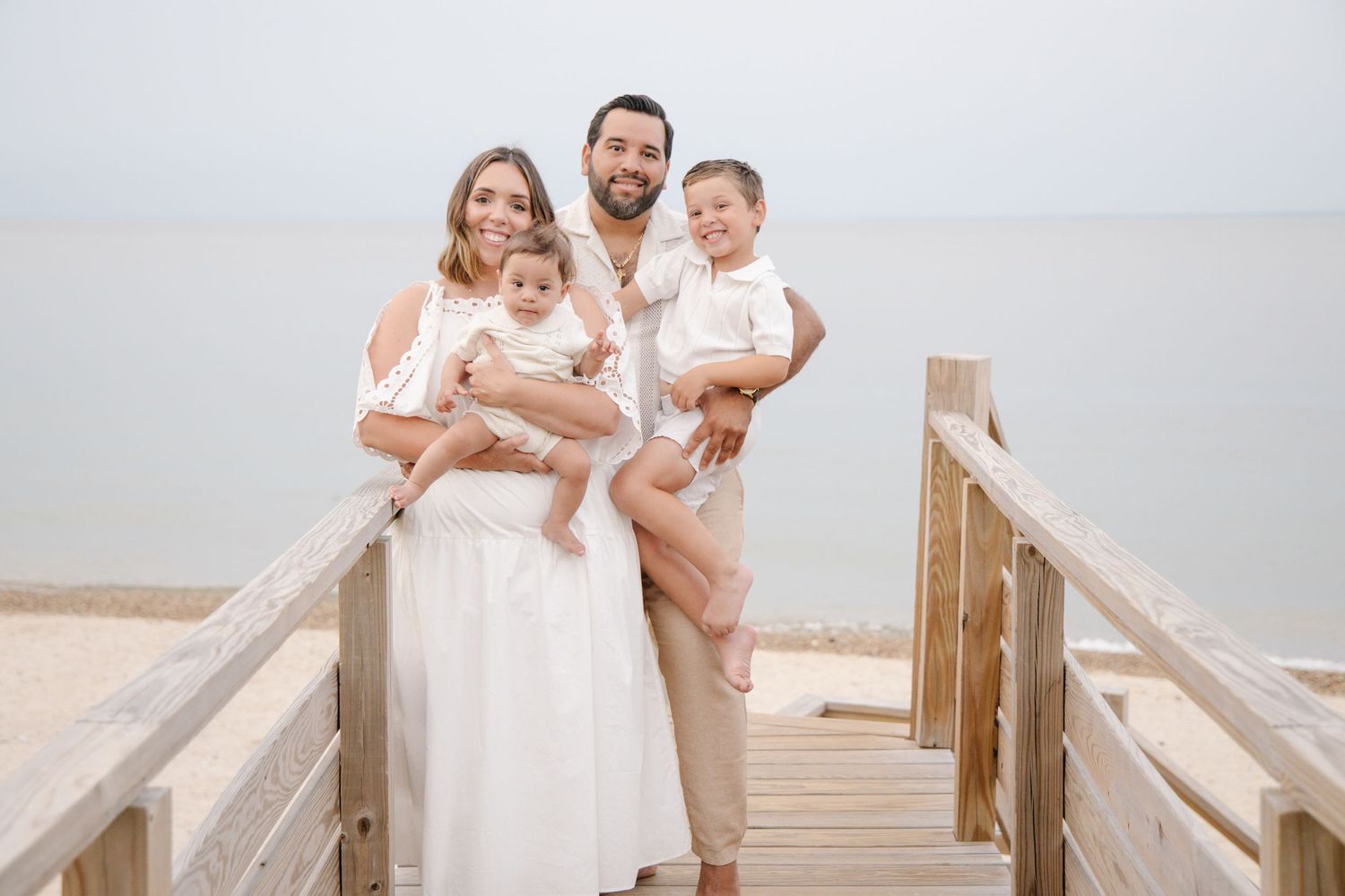 Happy family of four in white outfits posing on a wooden beach boardwalk on a calm day.