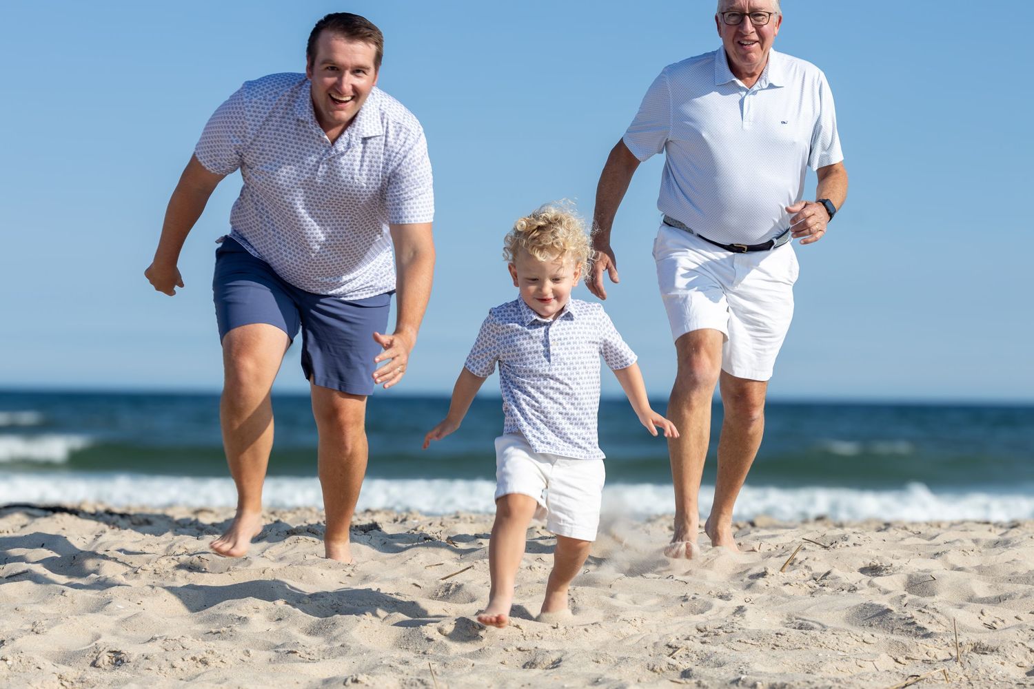 Two men and young blonde child running joyfully on sunny beach with ocean waves in background.