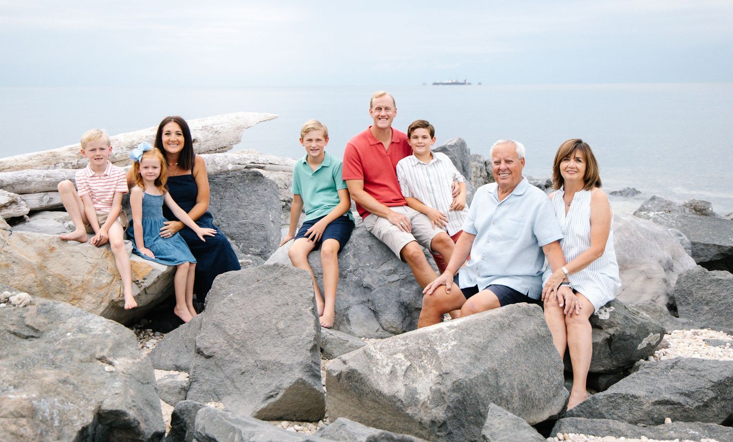 Multi-generational family of eight posing on large rocks by calm lake on a sunny day.