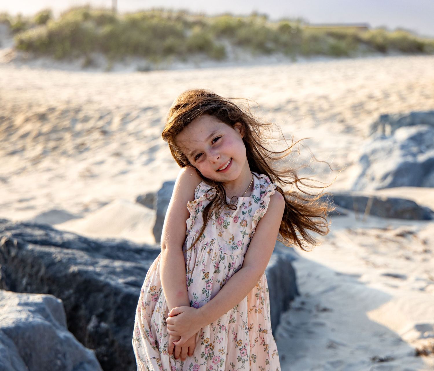 A young girl in a floral dress smiles on a sandy beach, her hair blowing in the wind near rocky shores. Hamptons photographer. beach session