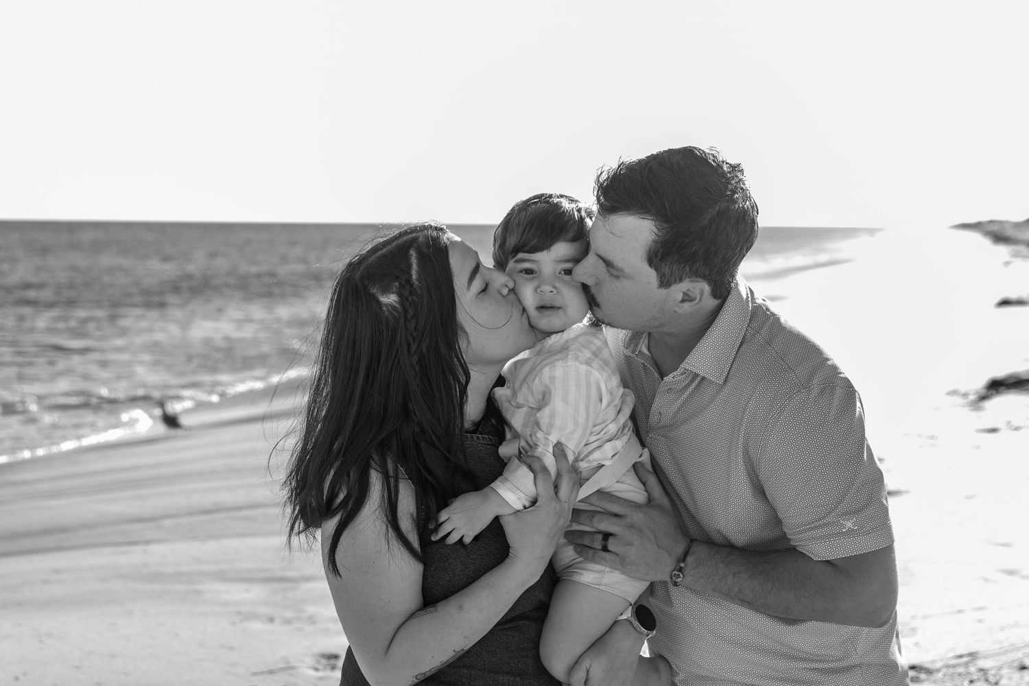 Black and white photo of parents kissing their baby on the cheeks at a sunny beach. Hamptons Beach. Family photographer. child session. NY