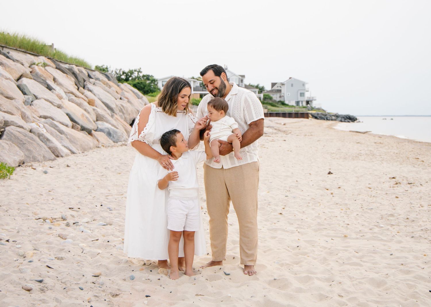Happy family of four posing together on a sandy beach with rocky shoreline backdrop.