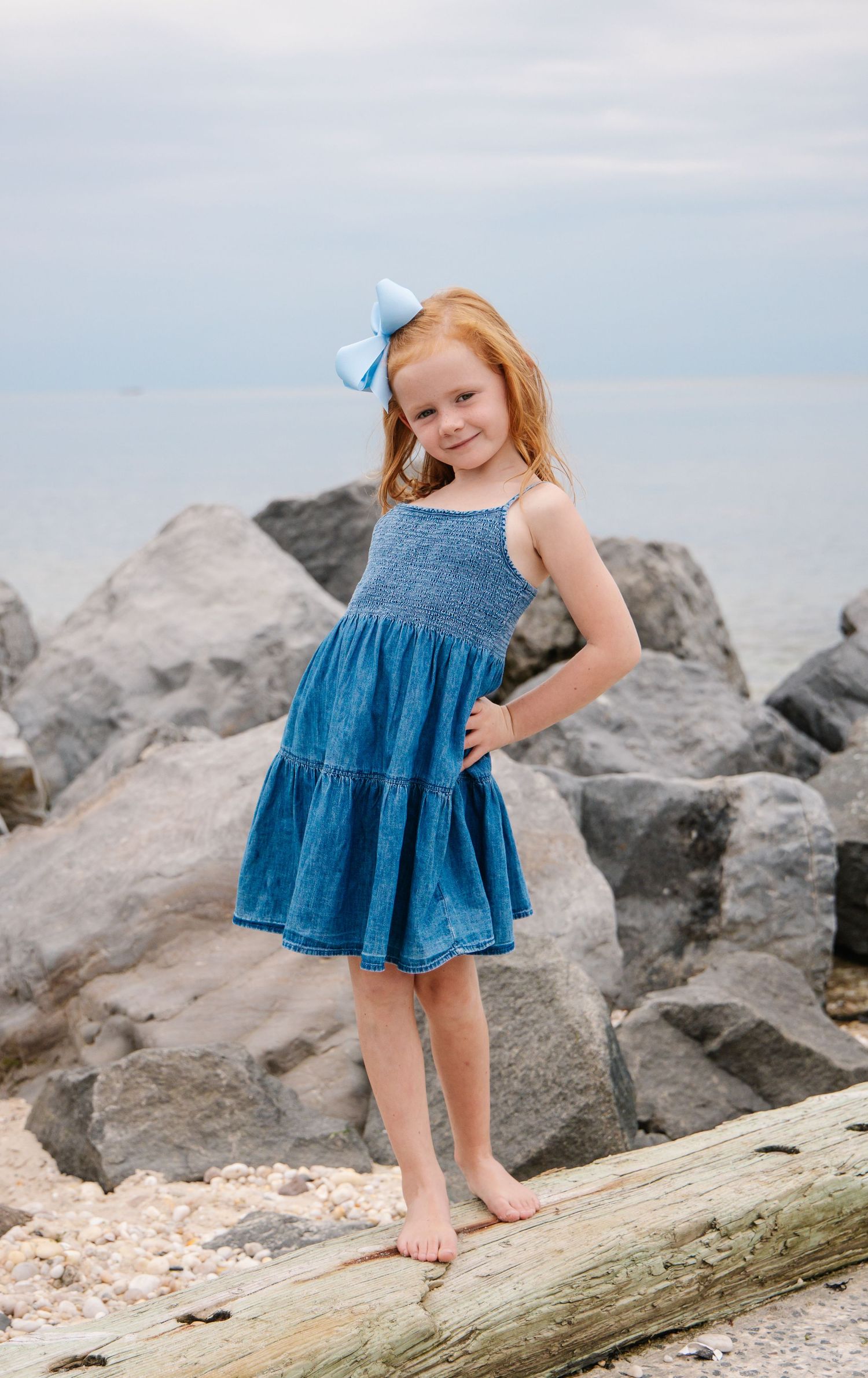 Redhead girl in a blue denim dress with a bow posing on rocks at the beach.