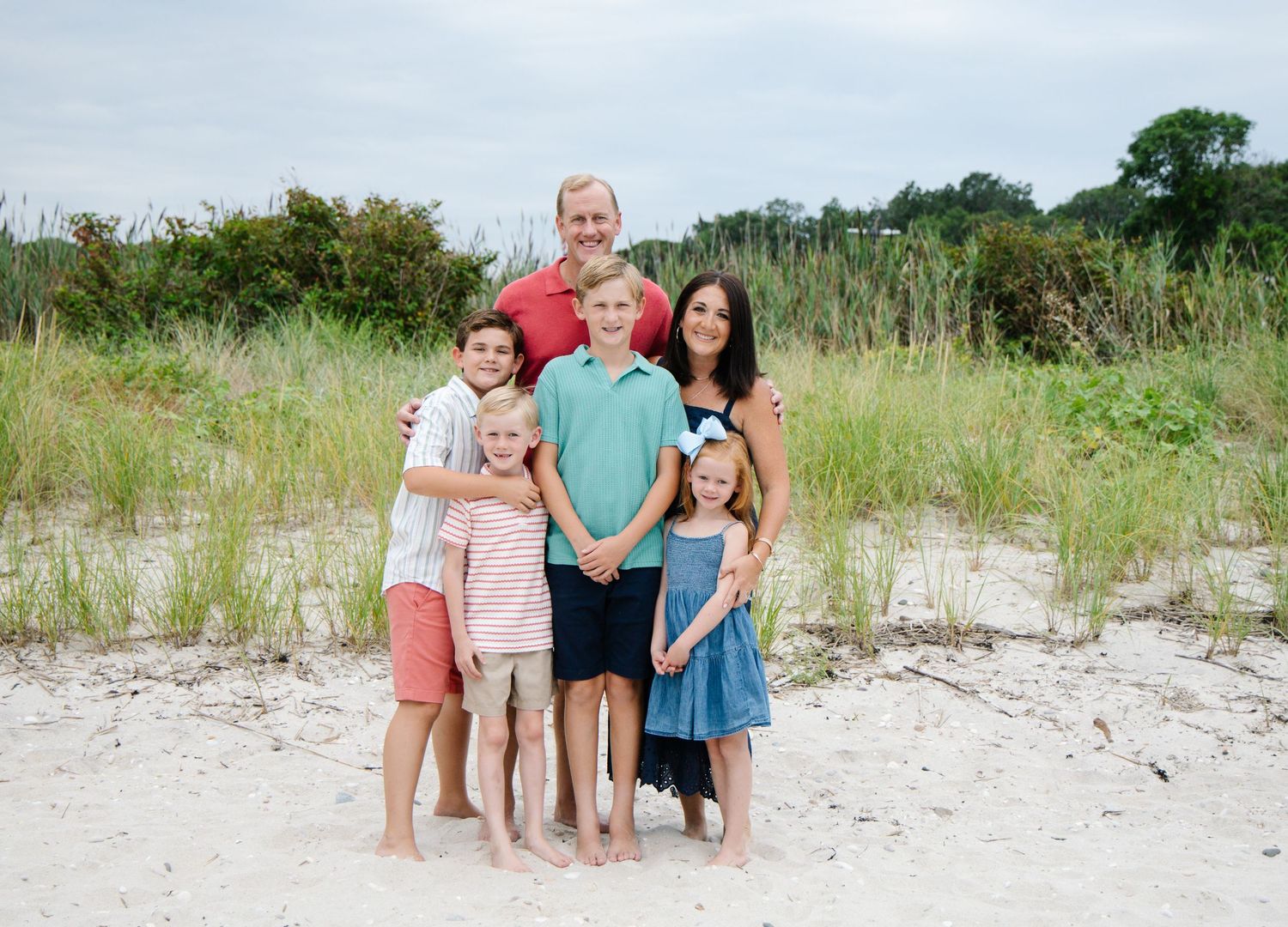 Happy family of six posing together on sandy beach with sea grass background.