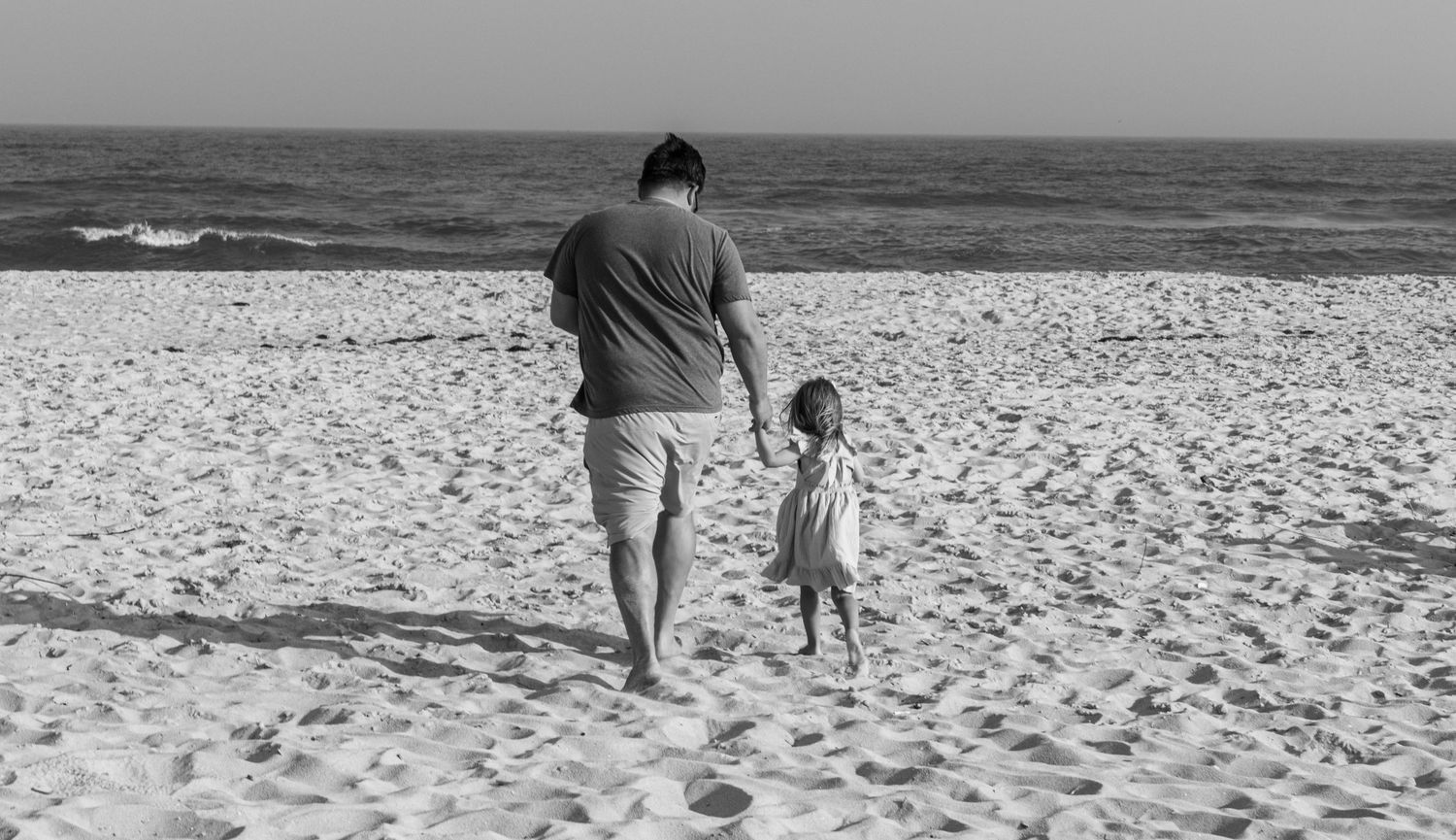 A father and young child walk hand-in-hand toward the ocean on a sandy beach in black and white.