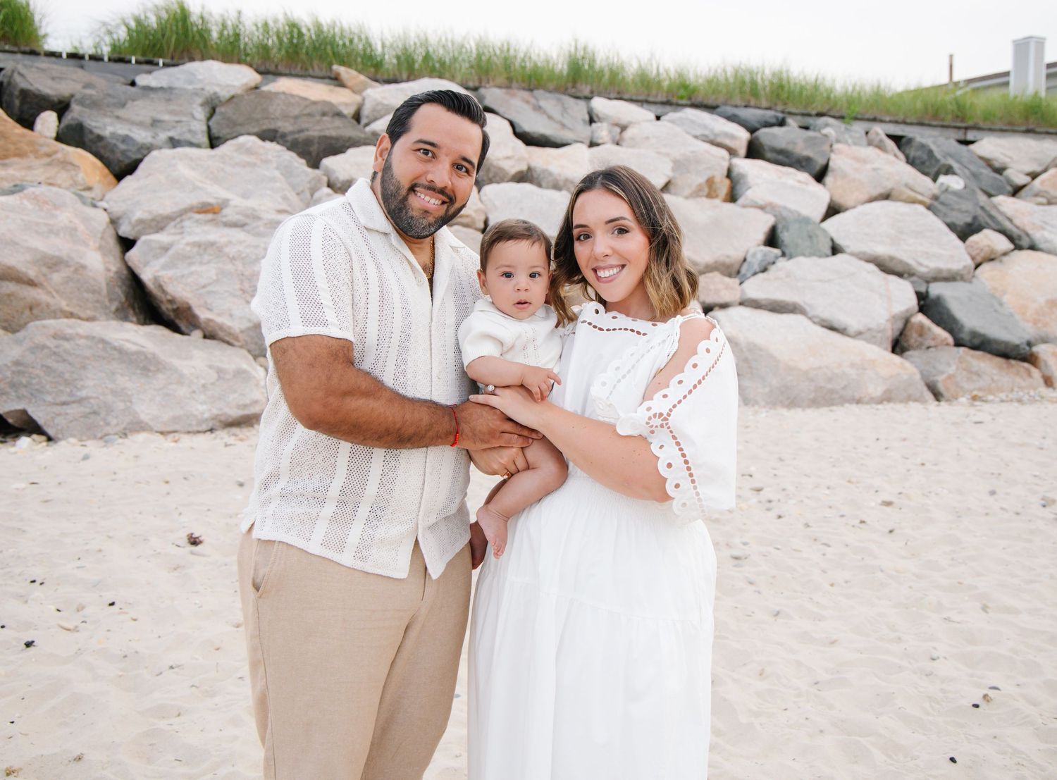 Couple in coordinating white and tan outfits holding baby girl on a sandy beach with rocks.