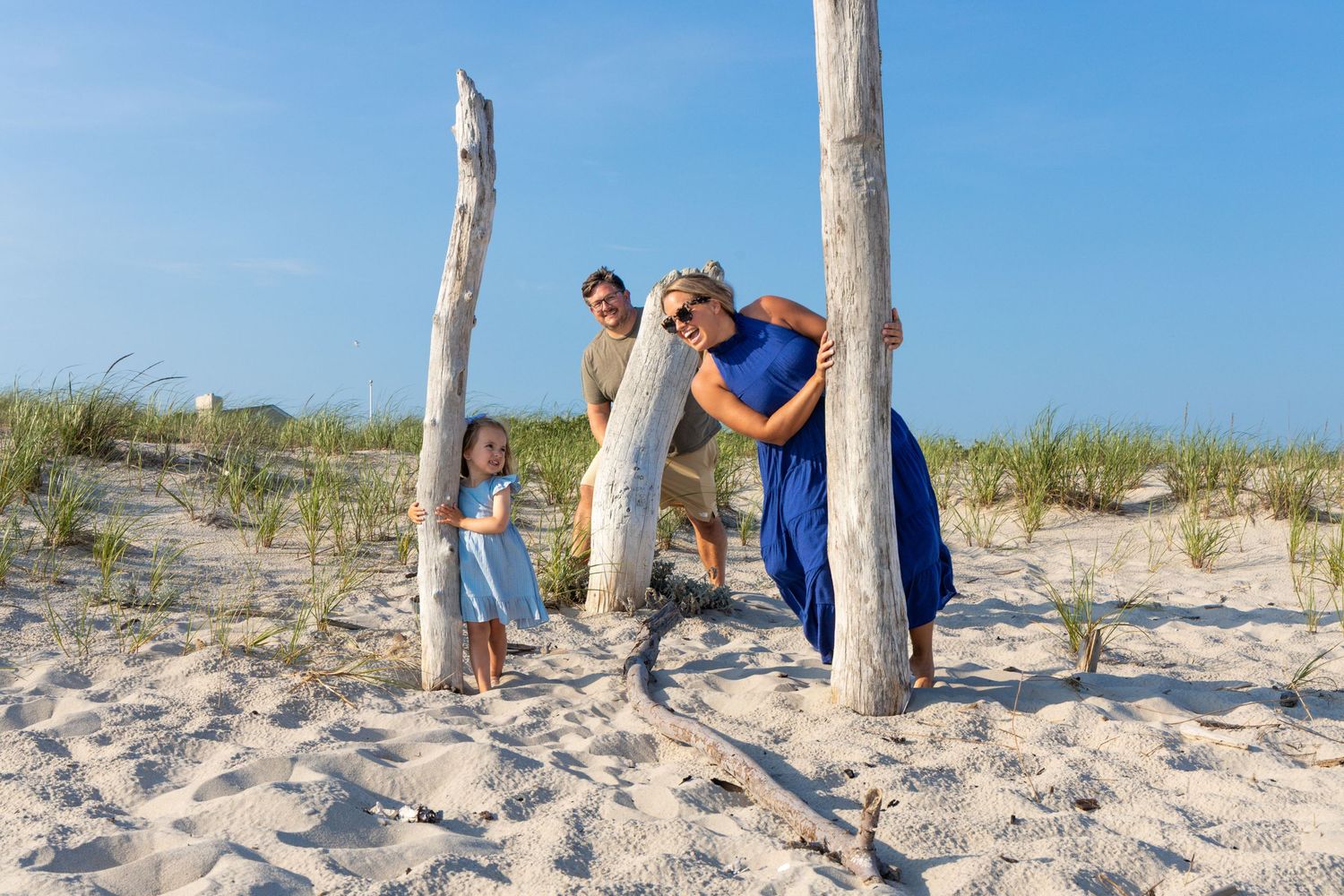 Happy family of four playing around driftwood poles on sunny sandy beach.