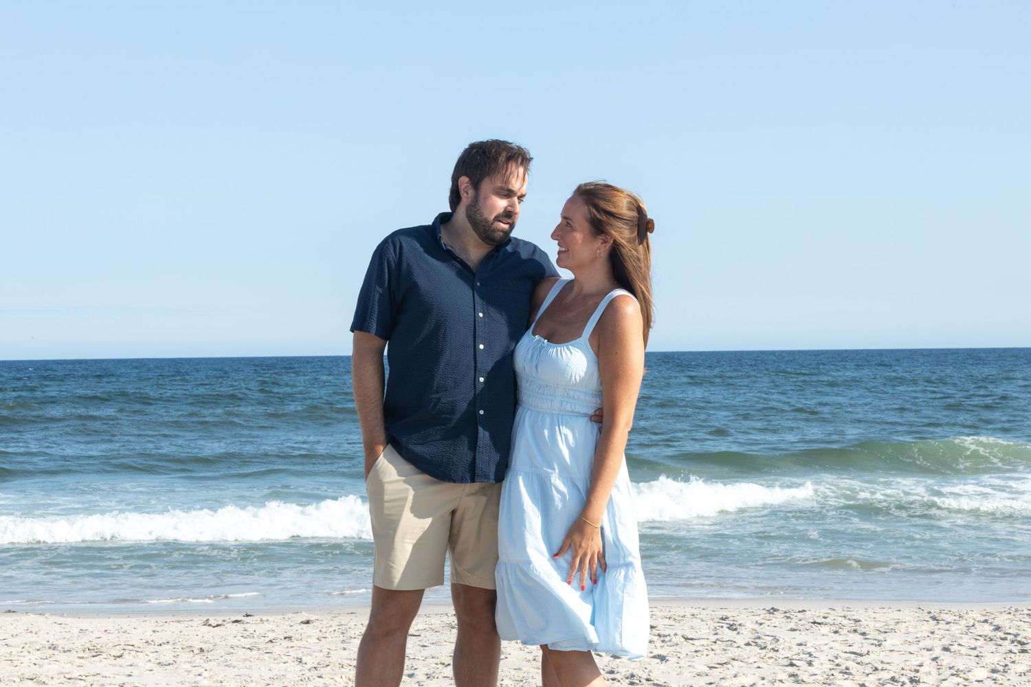Couple gazing at each other romantically on sandy beach, man in navy shirt and woman in blue sundress.
