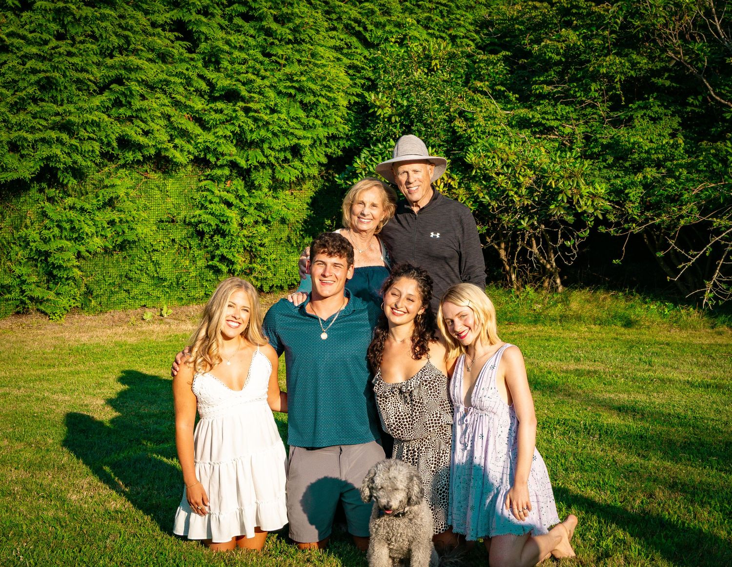Family of six posing outdoors on green lawn with gray poodle dog in golden evening light.