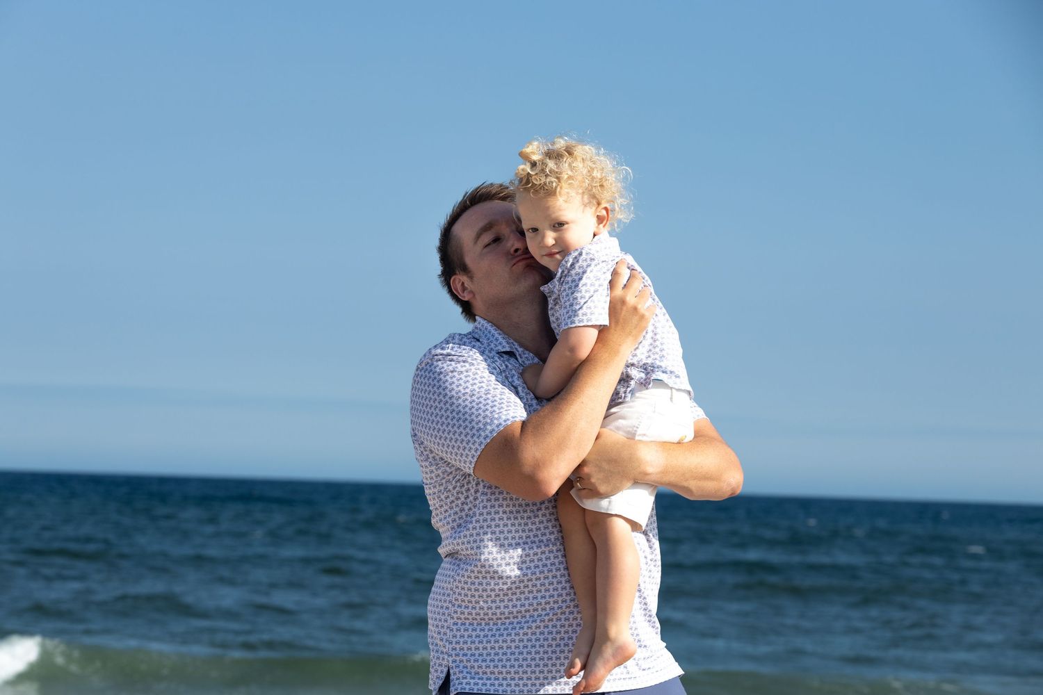 Father kissing curly-haired toddler on the cheek with blue ocean in the background.