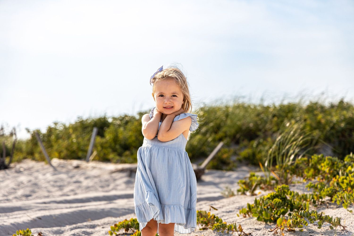 A young girl in a light blue dress poses on a sandy beach with green dunes behind her.