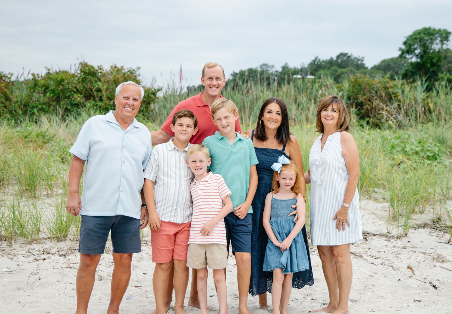 Extended family of eight posing on sandy beach with lush green dunes in background.