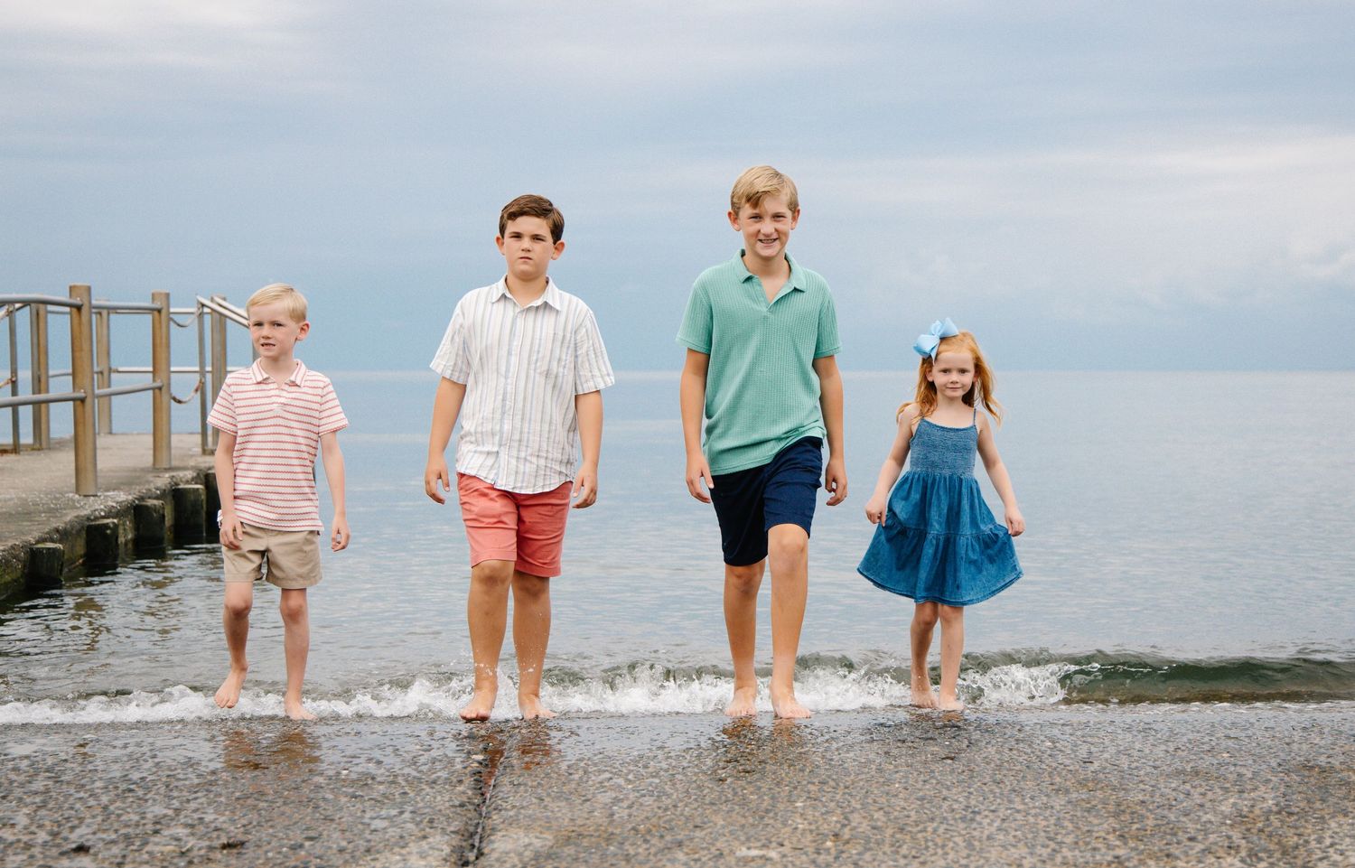 Four siblings walking along the shoreline at the beach on a cloudy day.