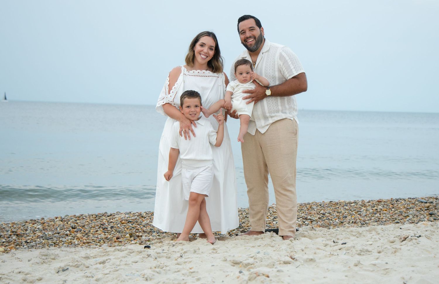 Family of four in white outfits posing together on a pebble beach with calm ocean behind.