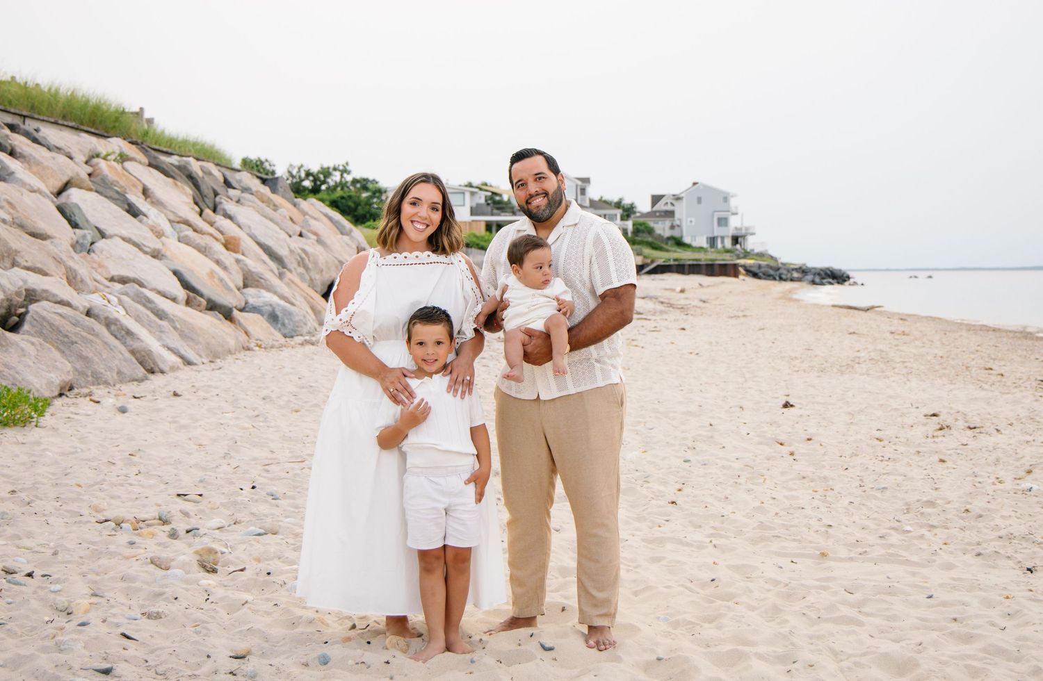 Family of four in white outfits posing together on a sandy beach with rocks.