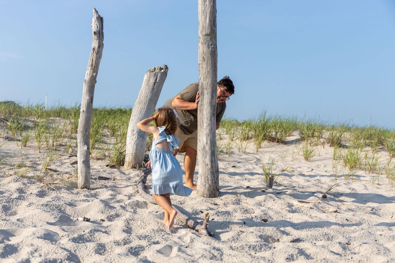 A father playfully chases his young daughter between driftwood posts on a sunny sandy beach.