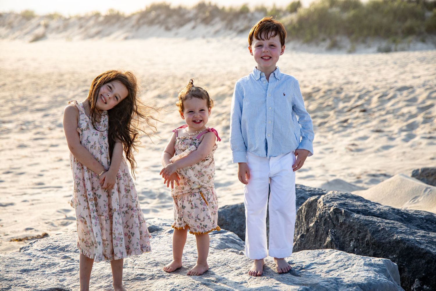 Three children pose on rocky beach at sunset, two girls in floral dresses and a boy in blue shirt with white pants. family photographer.