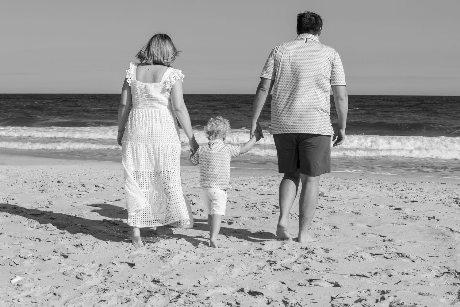Black and white rear view of couple walking on beach holding hands with small child between them.