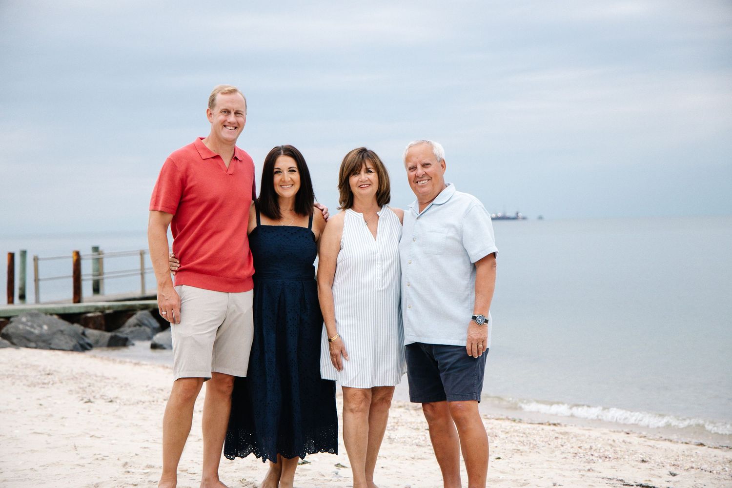 Two couples smile together on a rocky beach with calm ocean waters in the background.