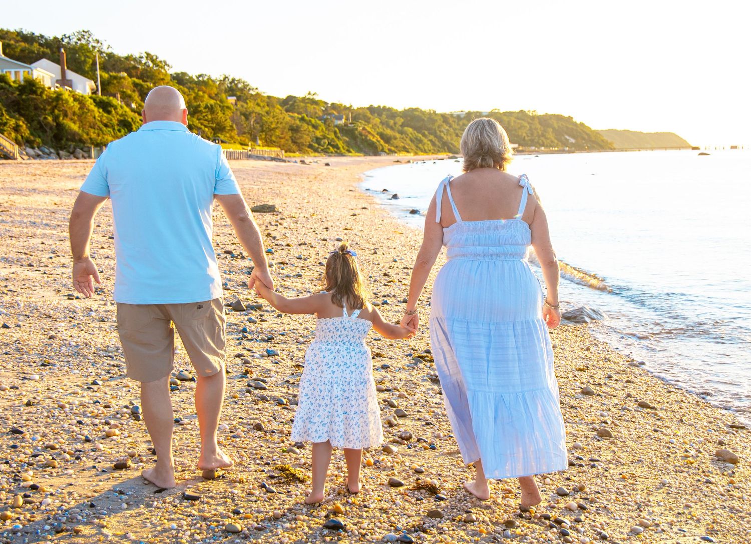 Family of three walking hand-in-hand on a sunny beach at golden hour, viewed from behind. north fork photographer. golden hour session