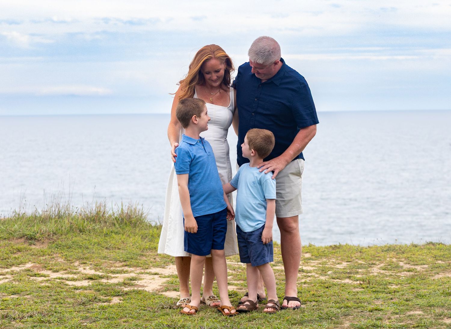 Family of four posing near ocean cliffs, two boys in blue shirts with parents on a sunny day.