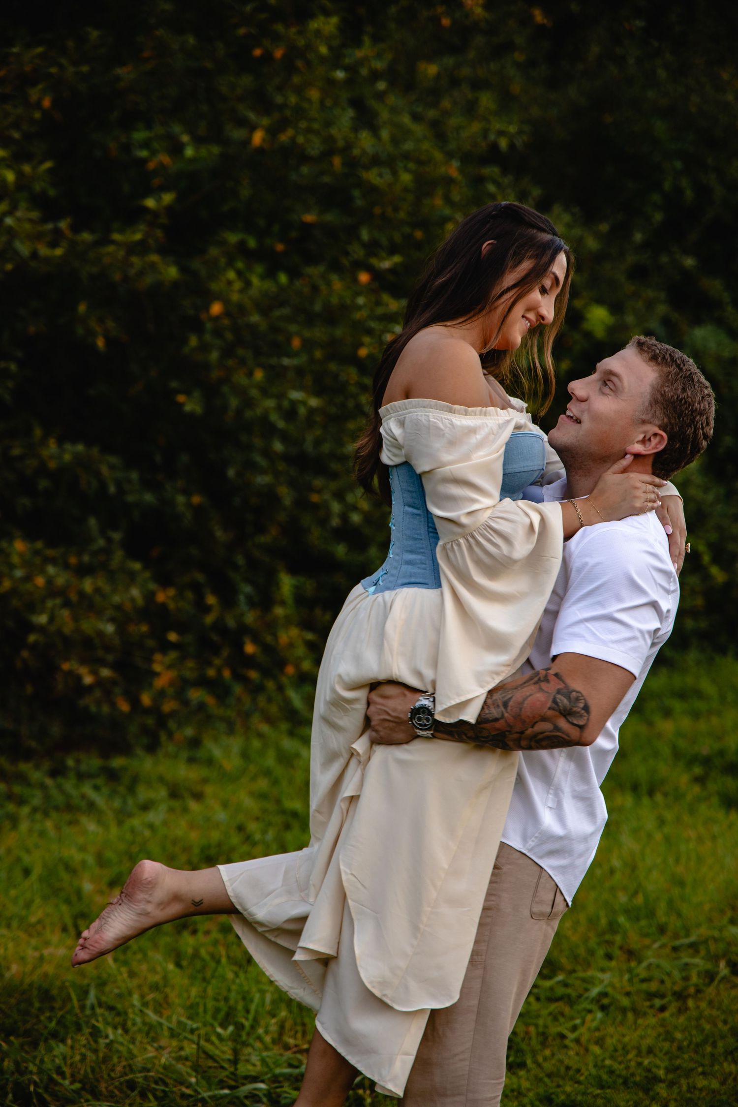 A man lifts and embraces a smiling woman in a blue off-shoulder dress outdoors near lush green trees. Engagement photographer. couples photo