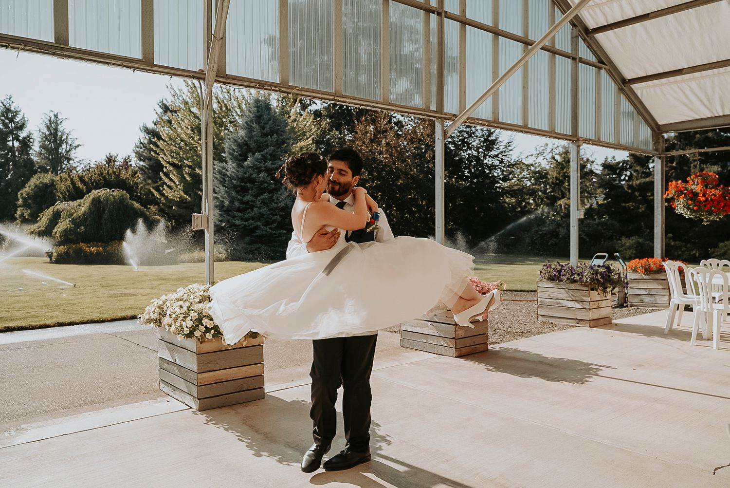 Groom carries bride in white dress under covered outdoor venue.