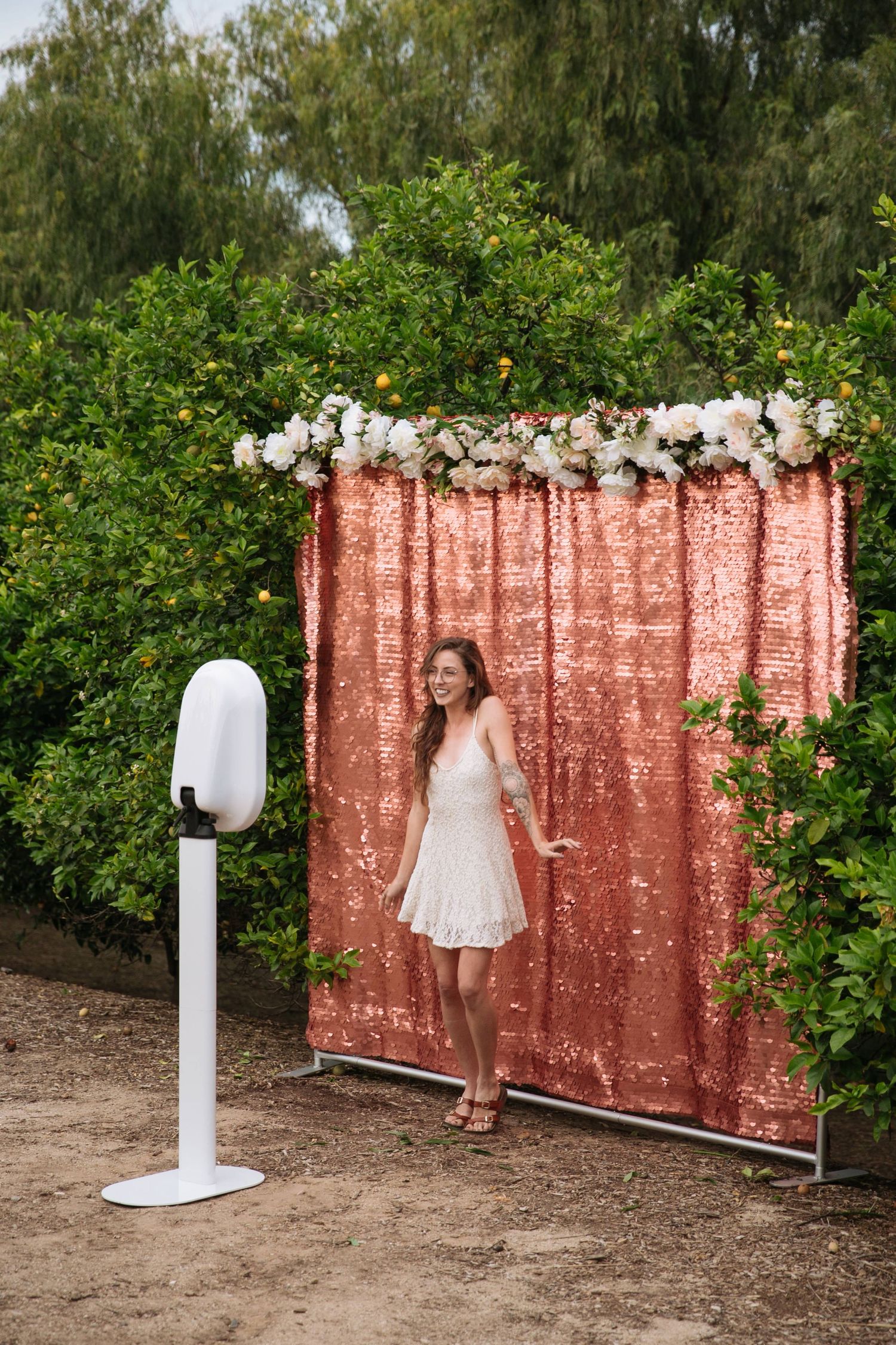 Photo booth setup with rose gold sequin backdrop and white floral decorations outdoors.