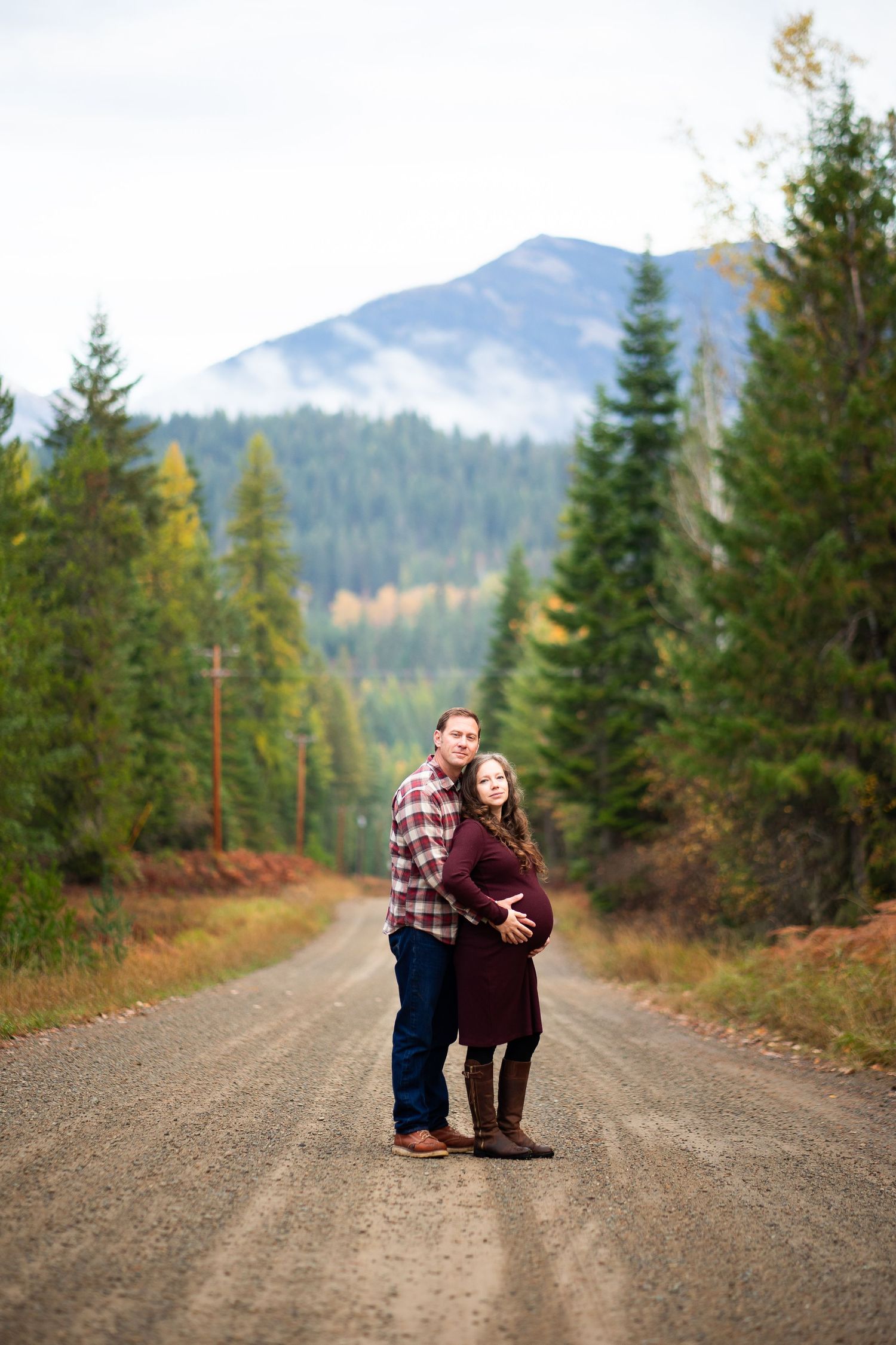 Expecting couple embracing on a scenic mountain dirt road surrounded by autumn evergreen trees.