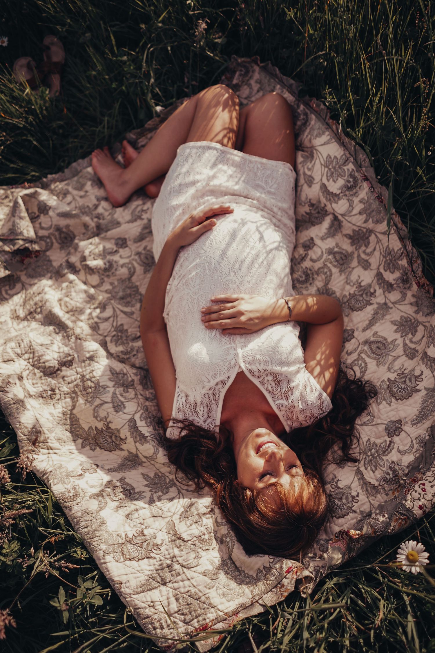 Pregnant woman in white lace dress lying on floral blanket in green grass with daisies.