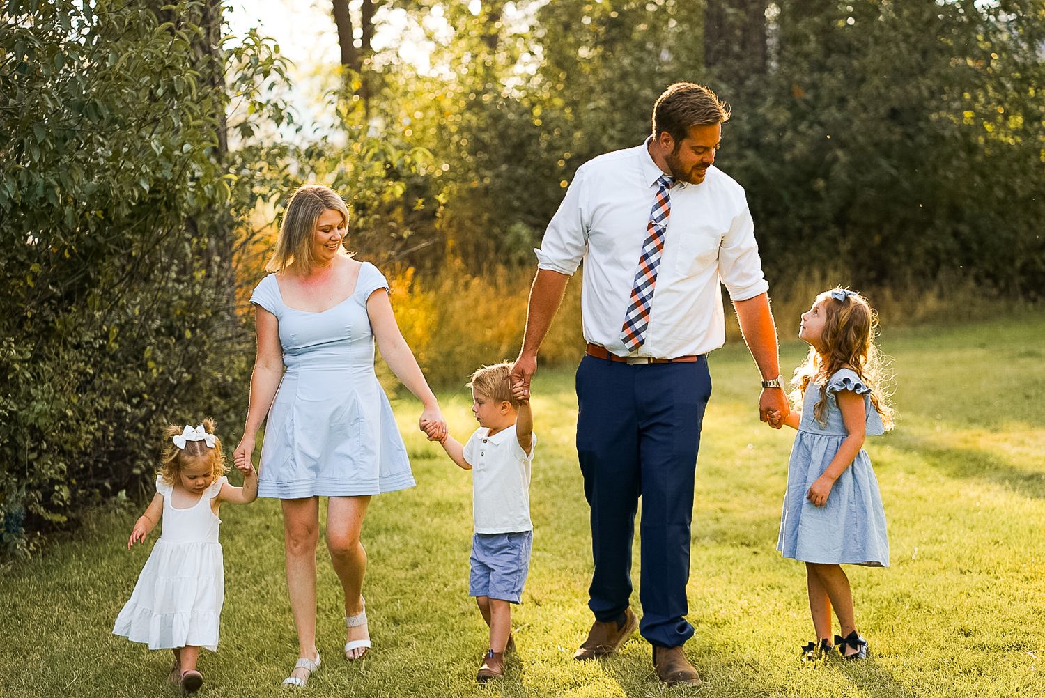 Happy family of five walking hand in hand through sunny green meadow at golden hour.