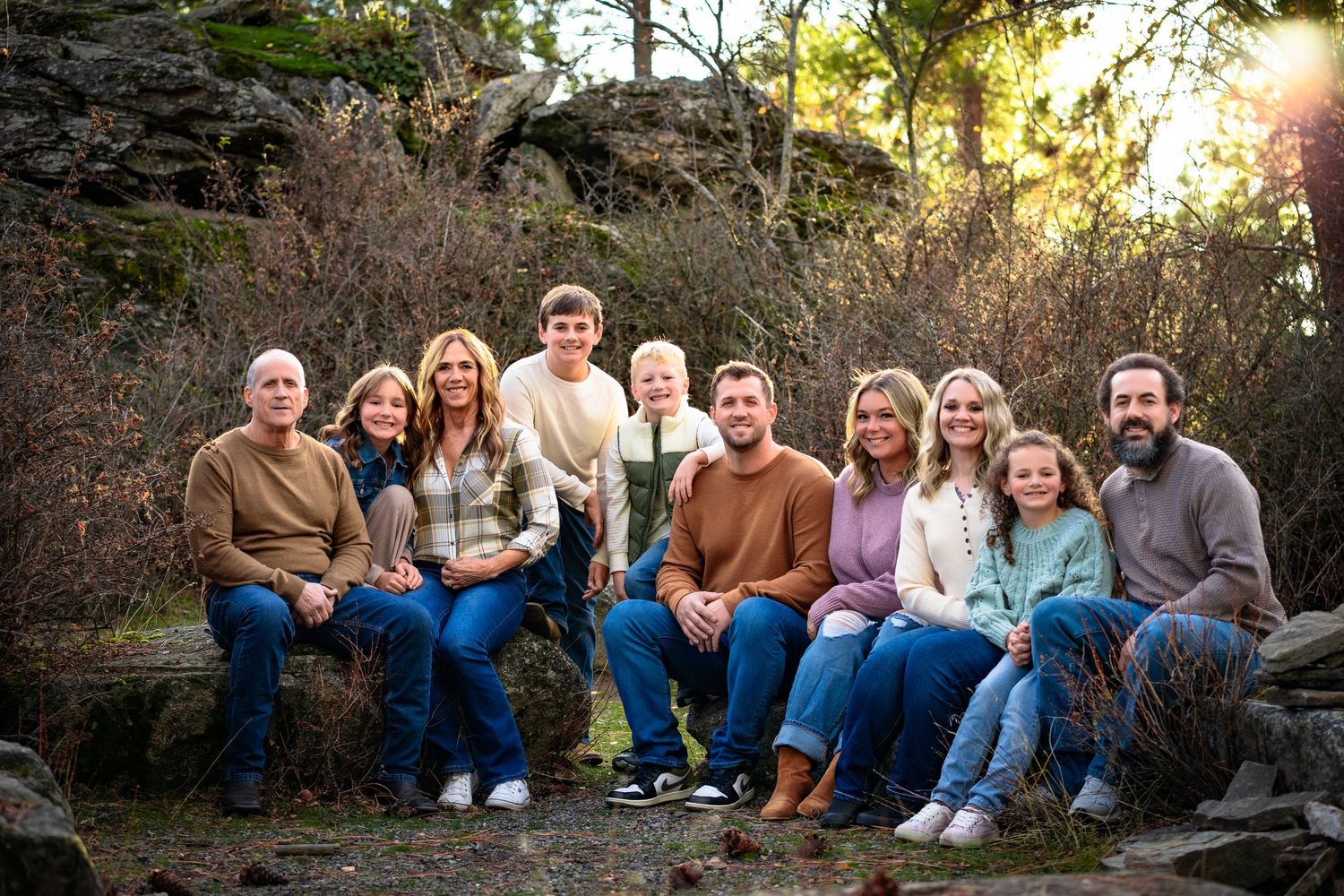 Extended family sitting together on stone wall in wooded outdoor setting during golden hour.