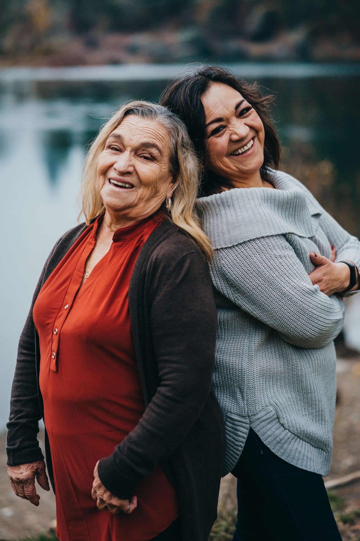 Two women laughing and embracing near a lake, one older in red dress and one in gray sweater.