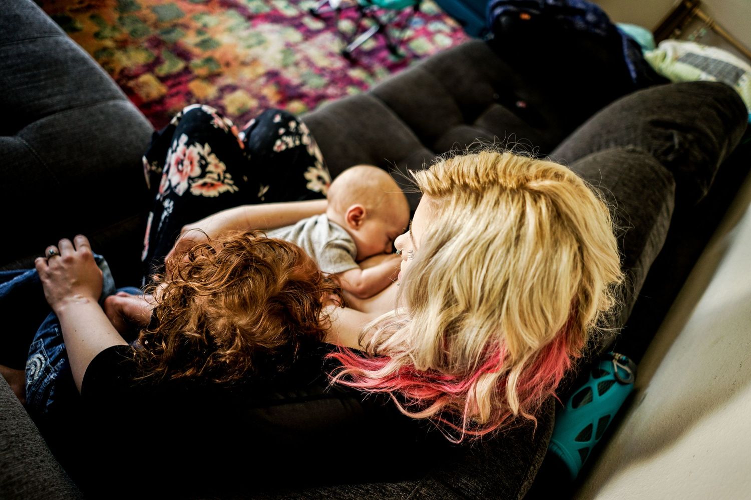 Blonde woman with pink highlights cuddling newborn baby on a dark couch indoors.