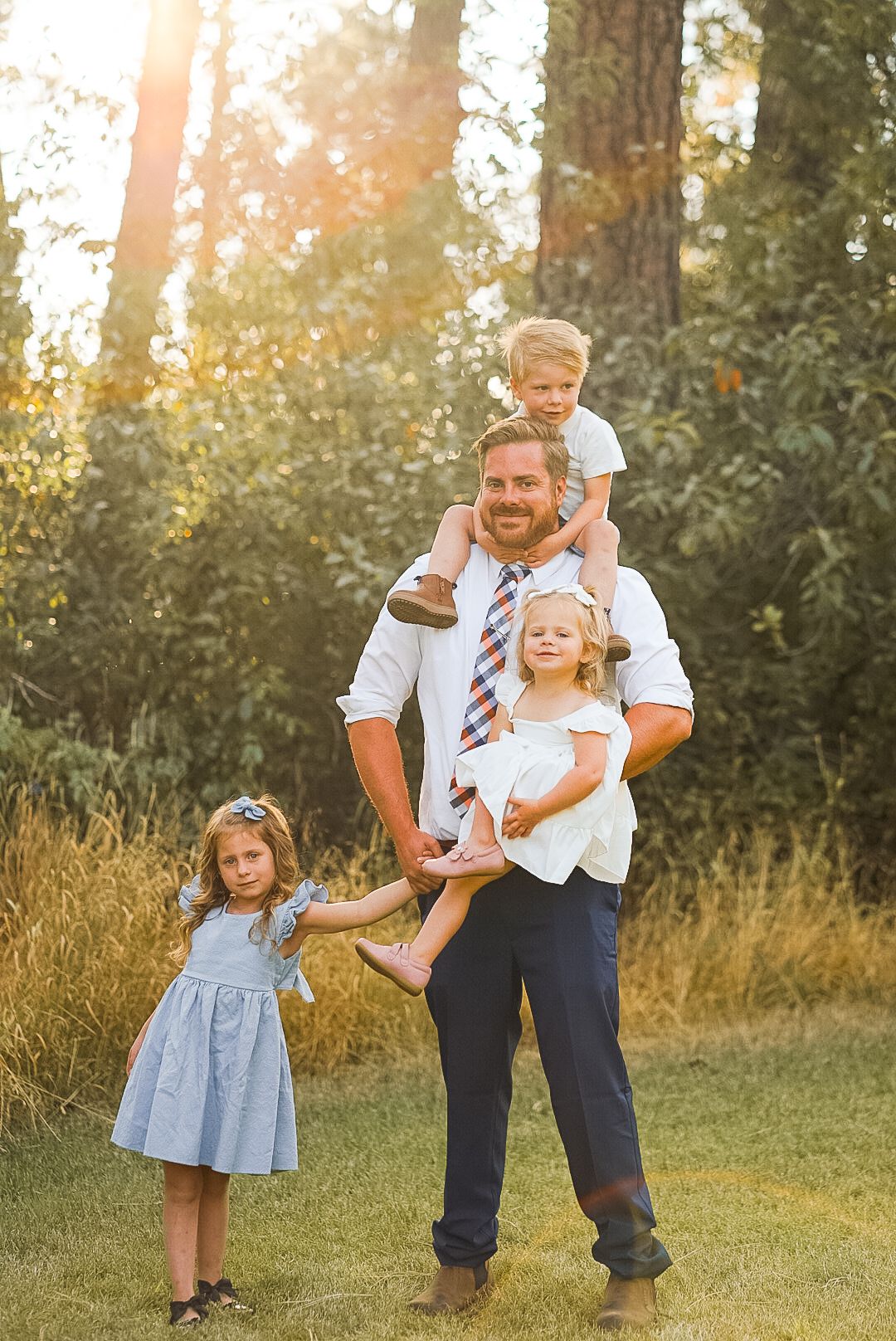 Father holding three young children in sunlit garden wearing white shirts and blue outfits.