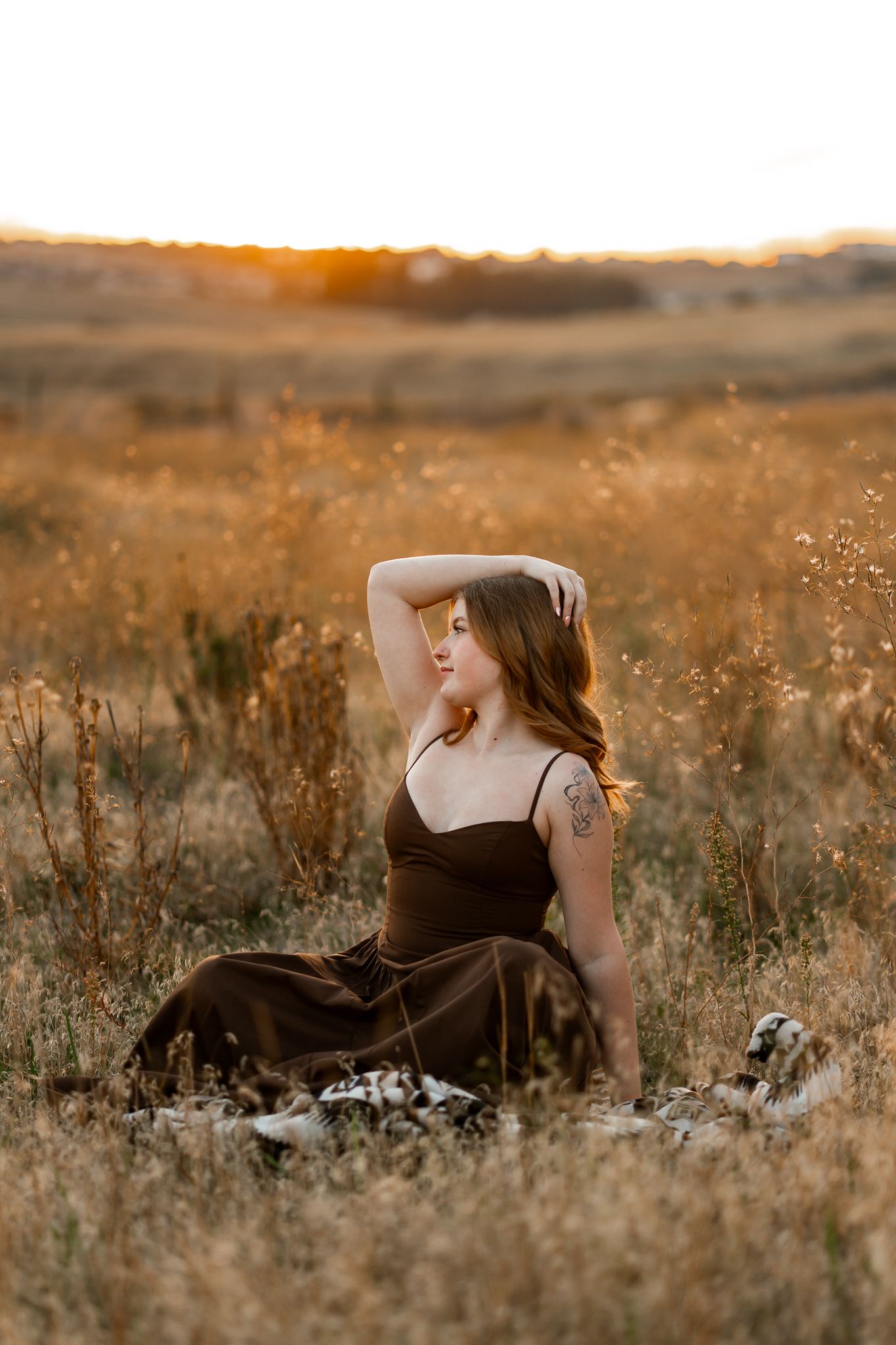 Woman in brown dress sitting in golden field at sunset with hand raised to head and tattoo visible on arm.