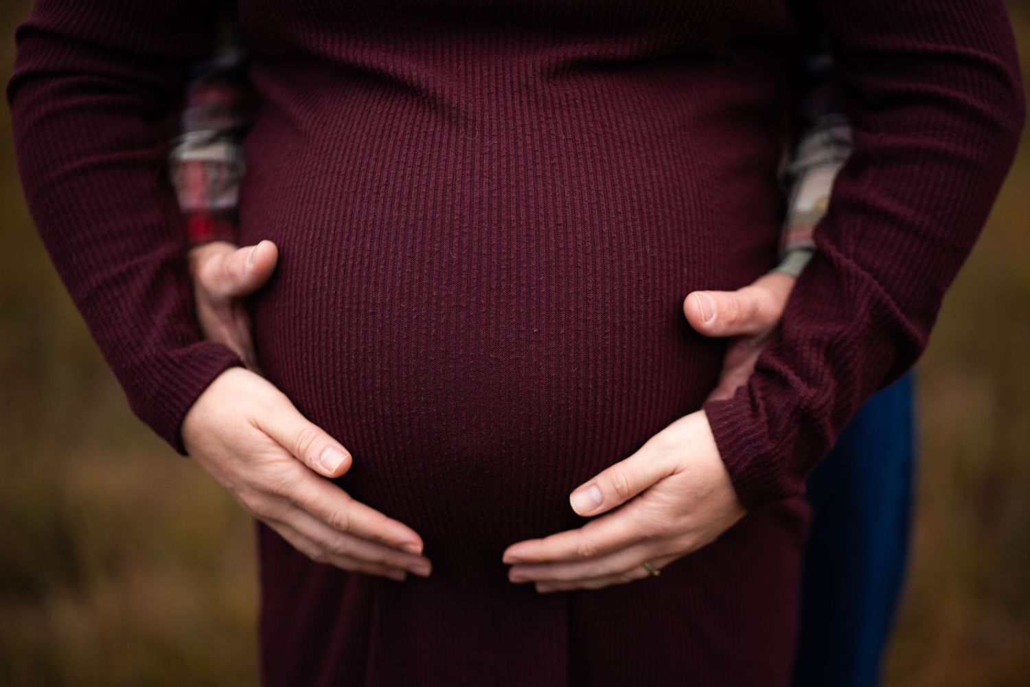 Close-up of pregnant belly being gently held by two pairs of hands in burgundy sweater.